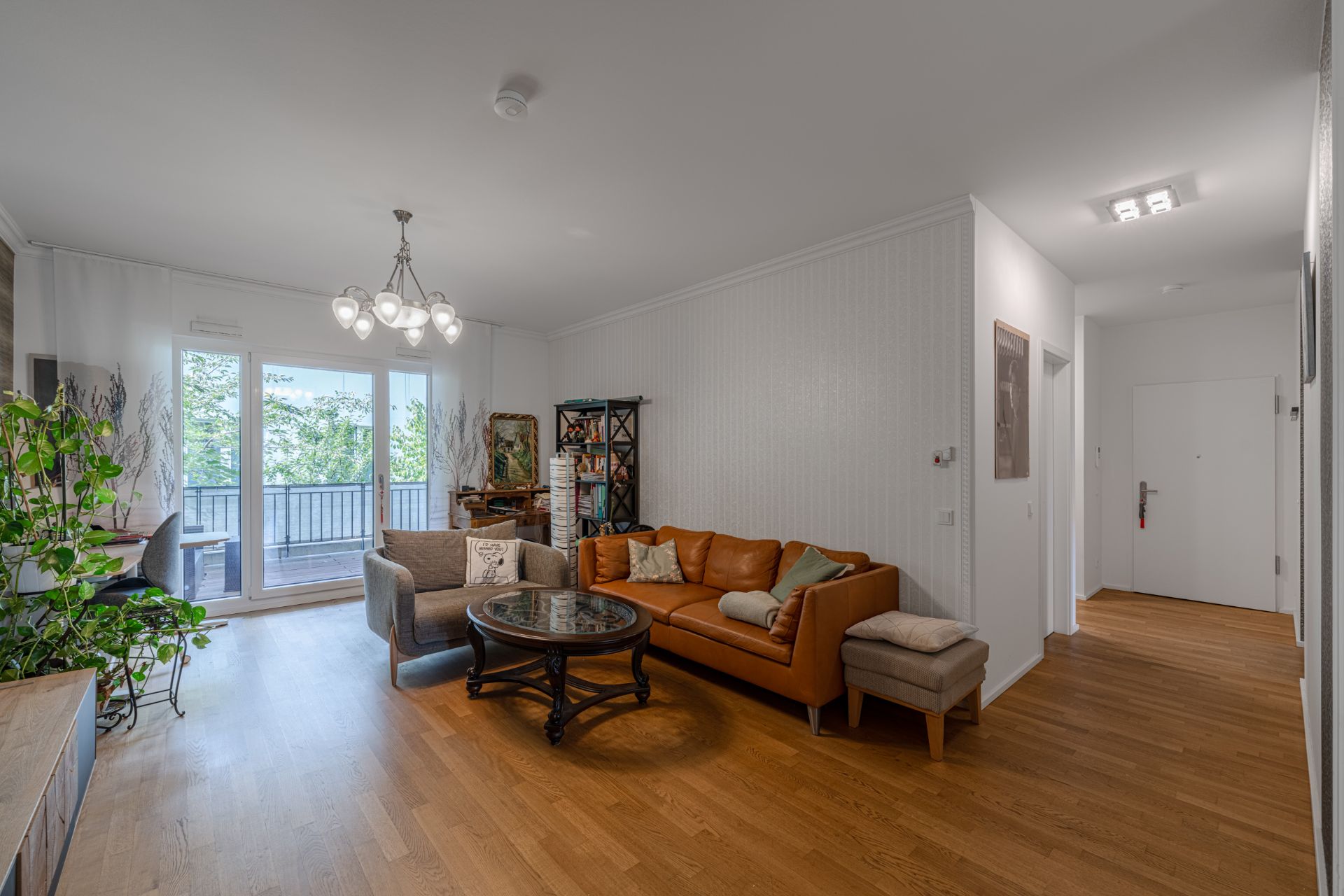Helles Wohnzimmer mit Sofa, Sessel, Glastisch, Regal und Zugang zum Balkon.