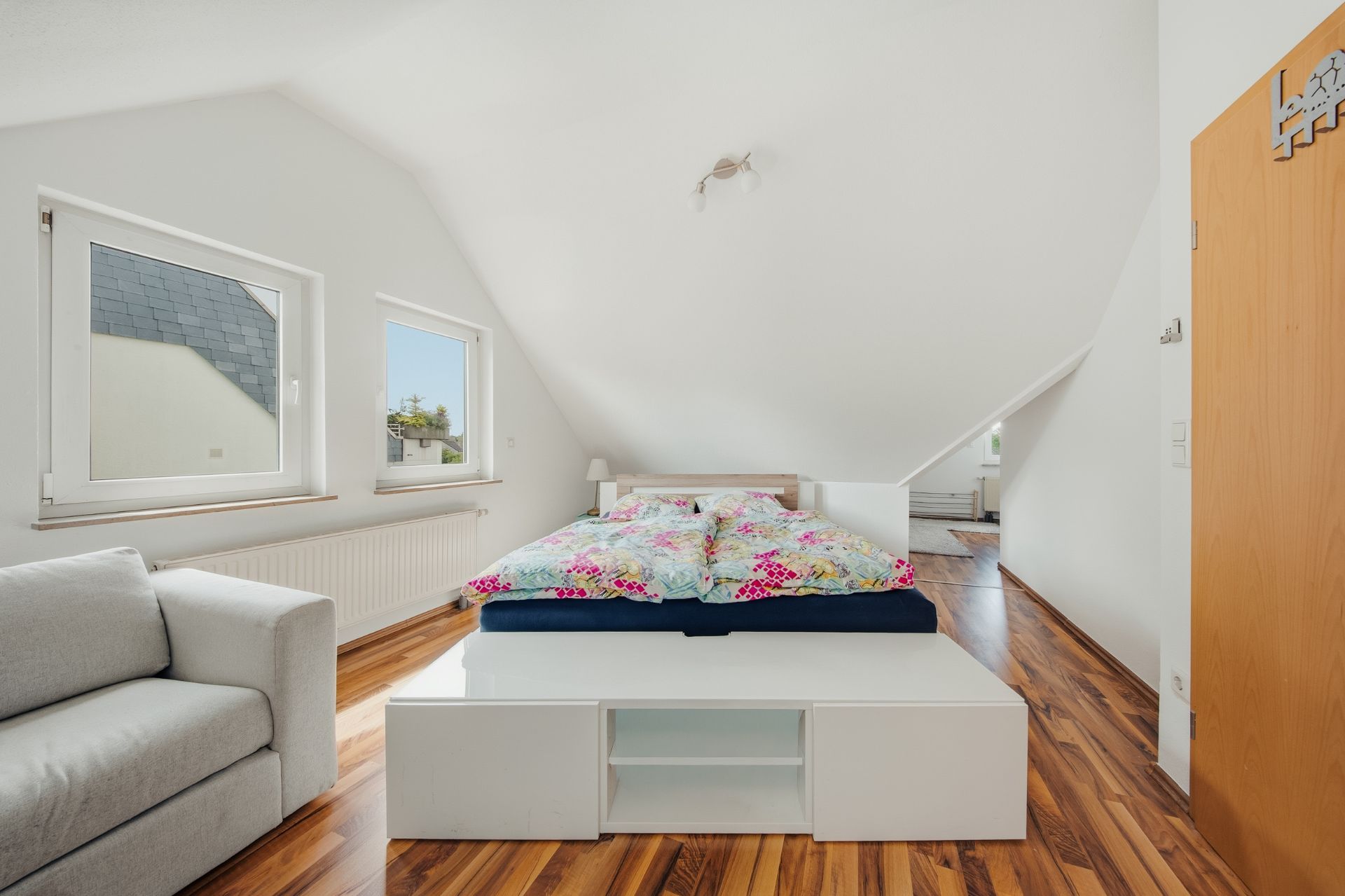 Helles Schlafzimmer mit Doppelbett, Sofa und zwei Fenstern unter Dachschrägen.