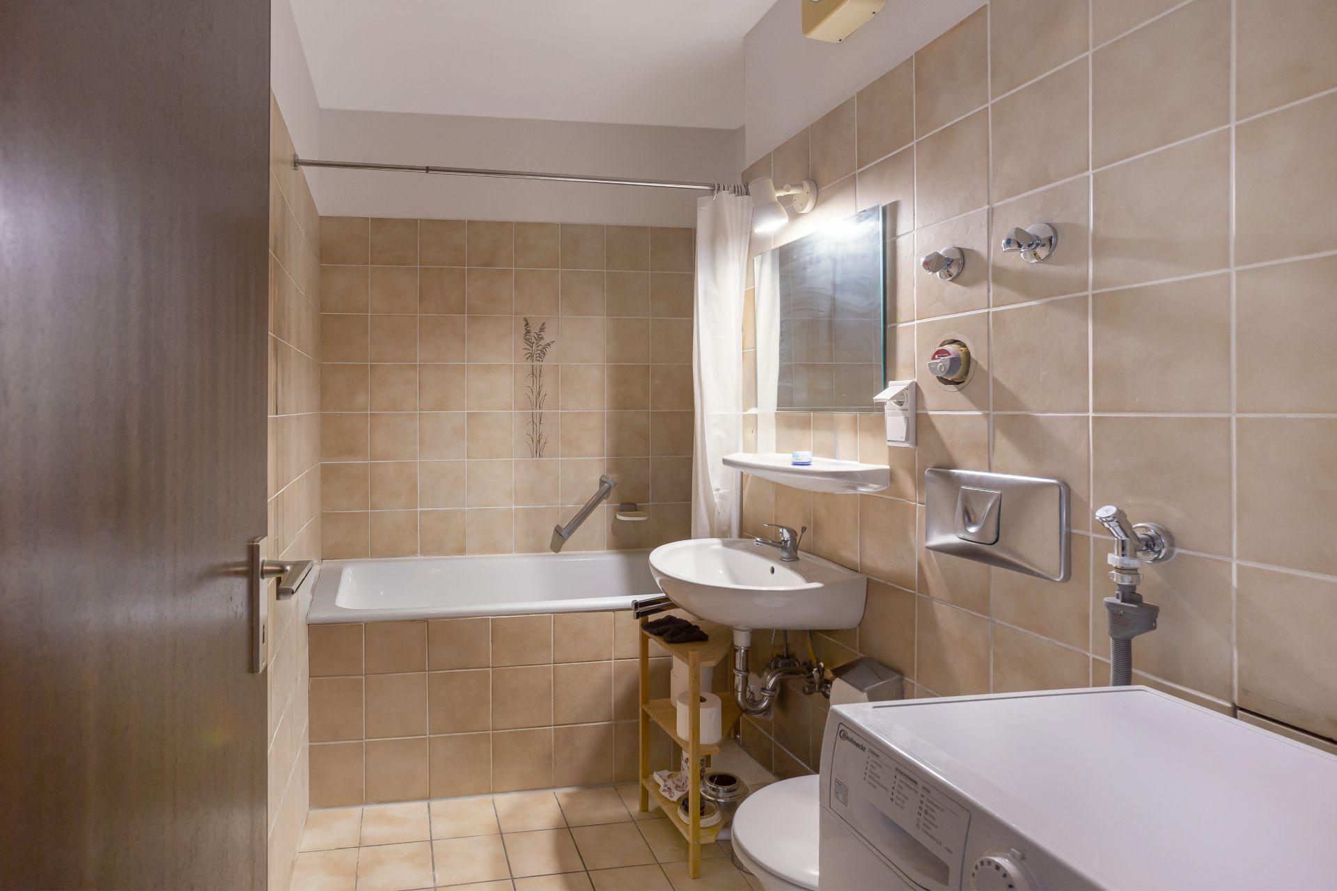 Kleines Badezimmer mit Badewanne, Waschbecken, Toilette, Waschmaschine und beigen Fliesen.