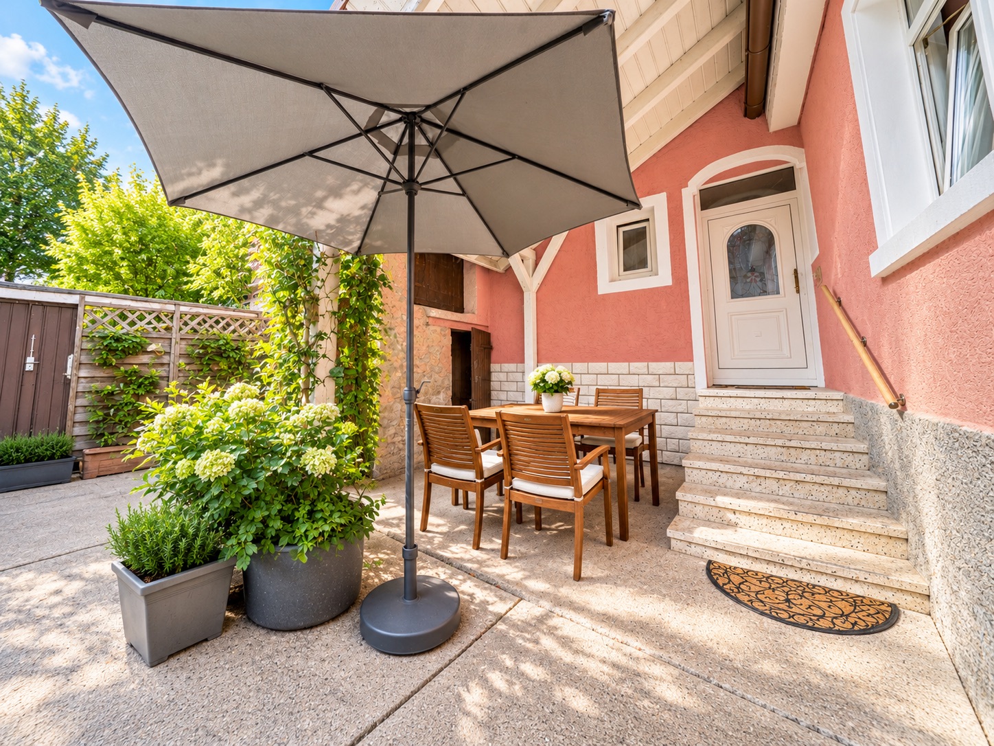 Sonnige Terrasse mit großem Sonnenschirm, Holztisch, Stühlen und Pflanzen neben einer Hauseingangstreppe.