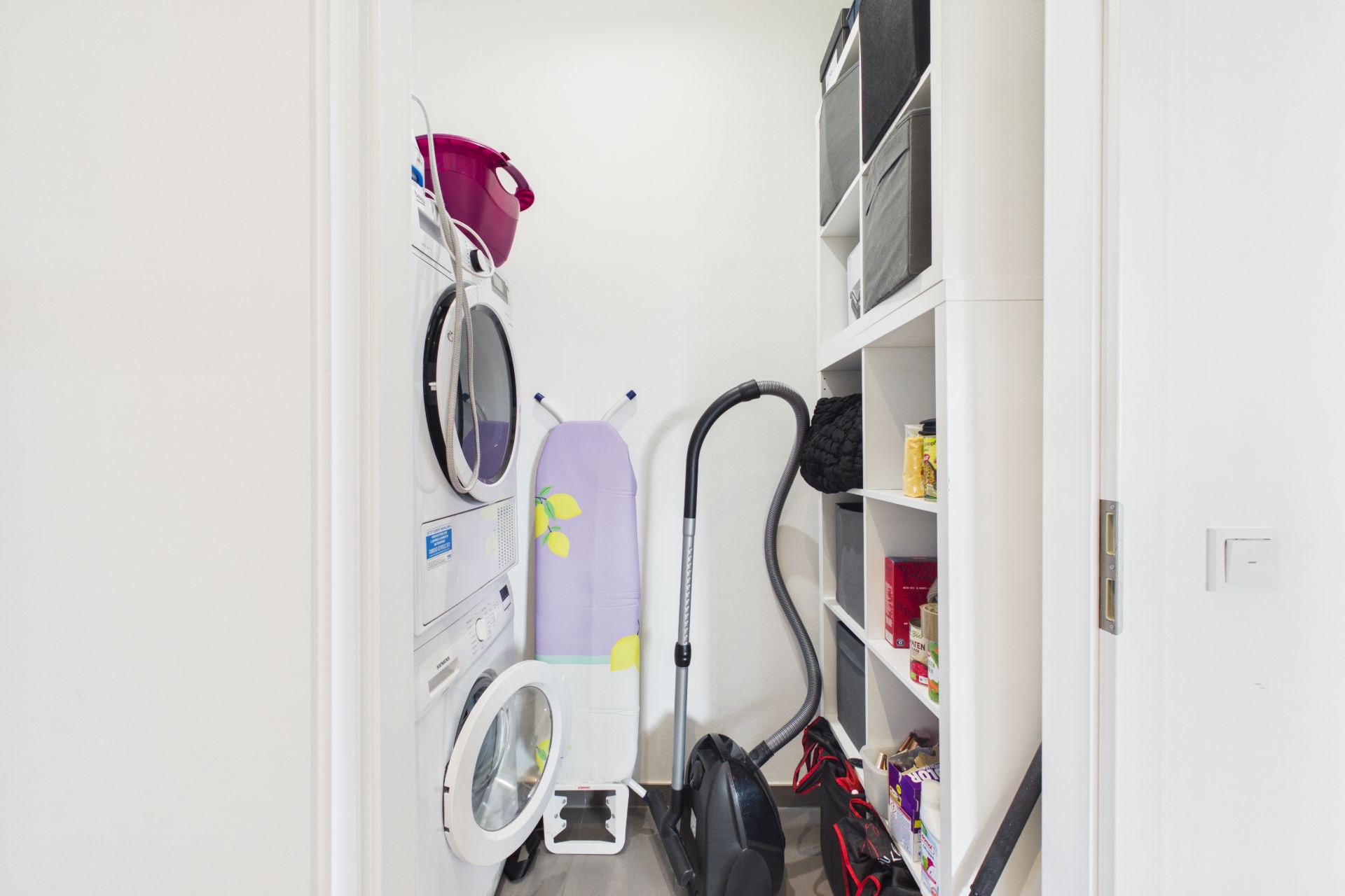 Hauswirtschaftsraum mit Waschmaschine, Trockner, Bügelbrett, Staubsauger und Regalen mit Aufbewahrungsboxen.