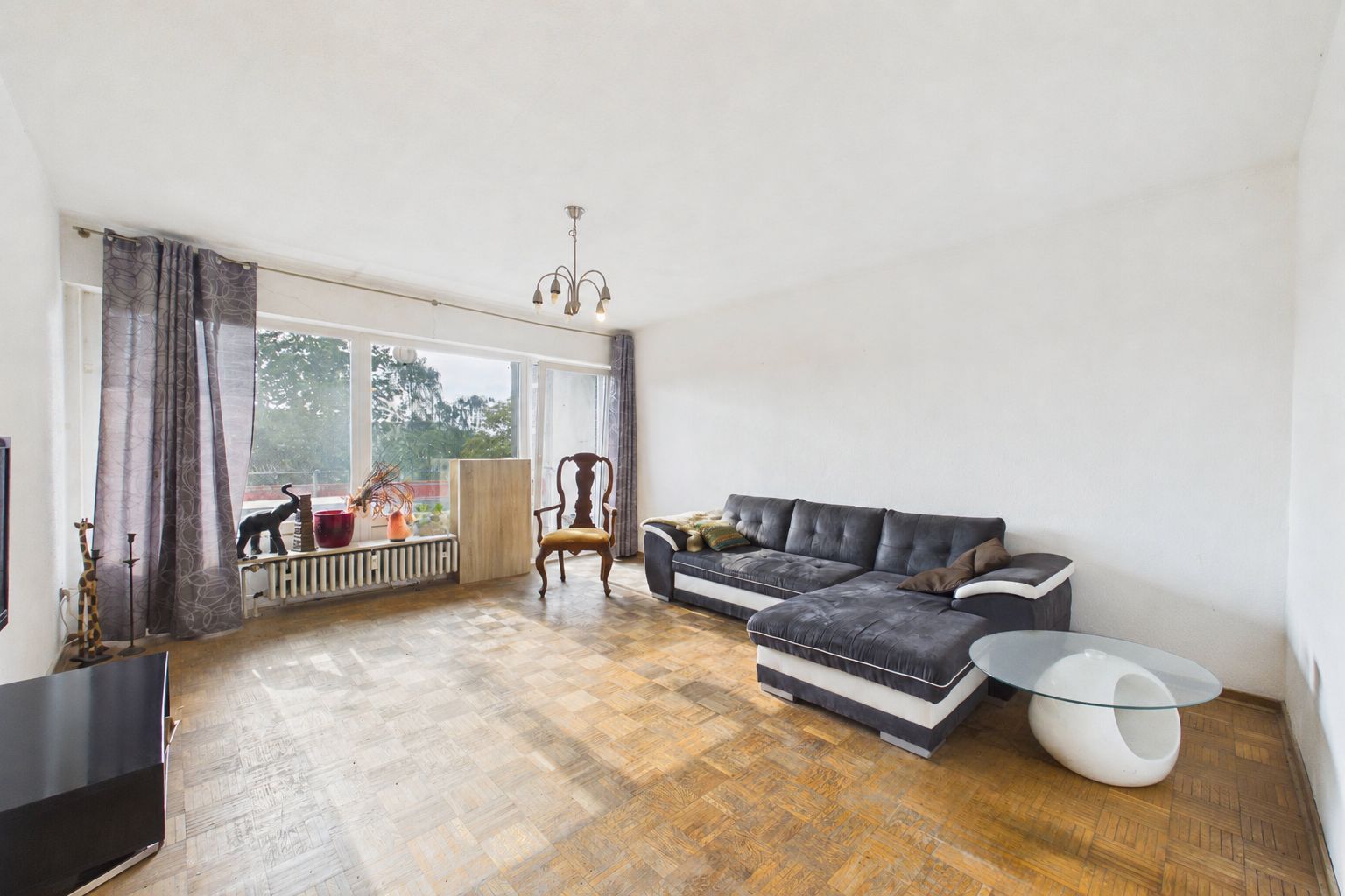 Wohnzimmer mit Ecksofa, Glastisch, Sessel, Fernseher und großen Fenstern mit Vorhängen.