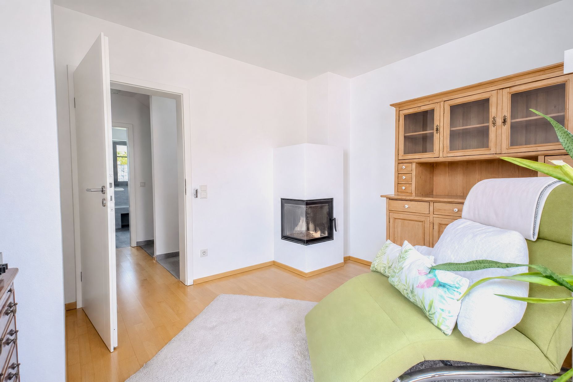 Helles Wohnzimmer mit grünem Sessel, Kamin und Holzvitrine auf Parkettboden.