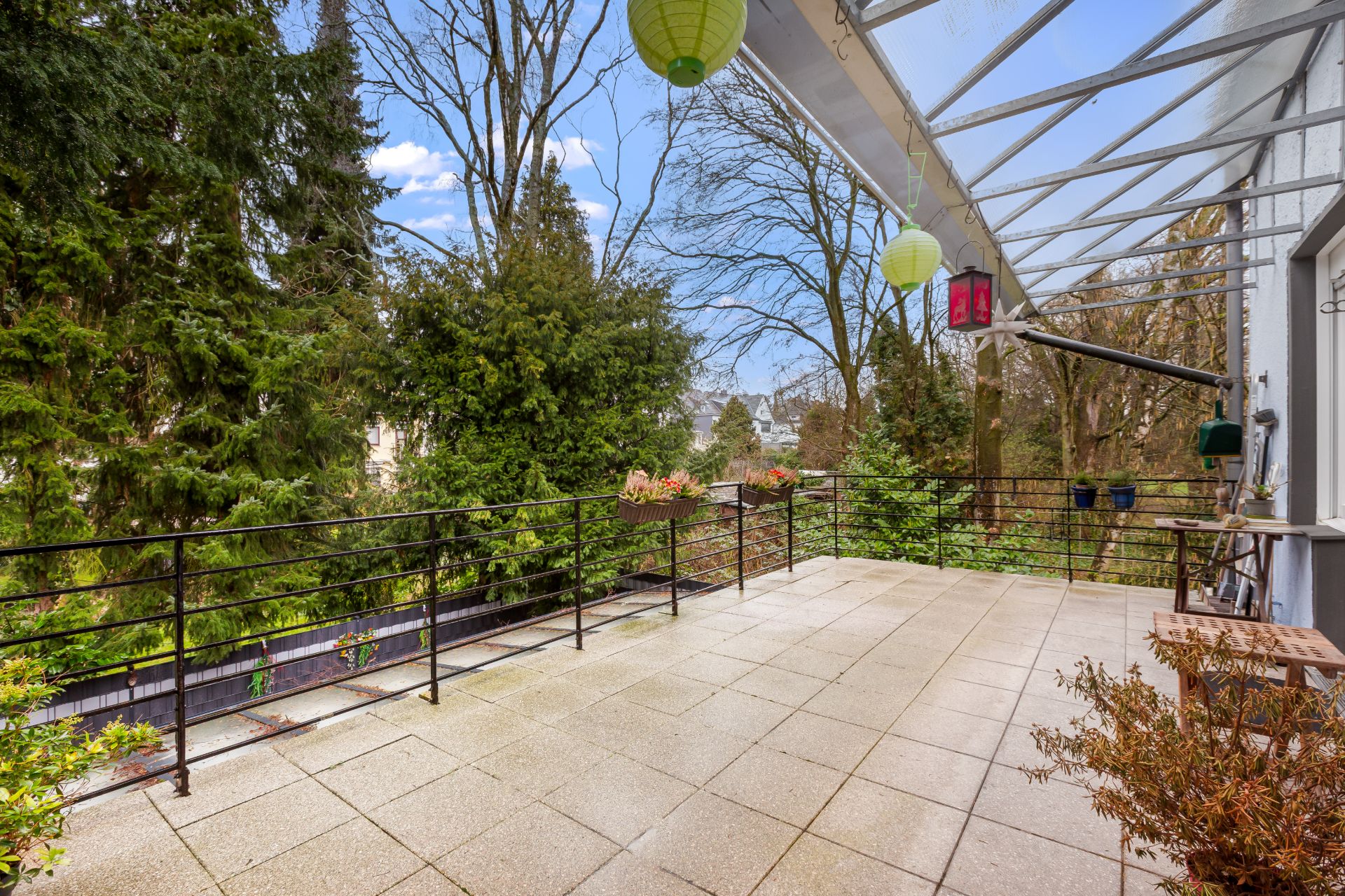 Große Terrasse mit Geländer, Pflanzen und Blick auf Bäume, überdacht mit Laternen.