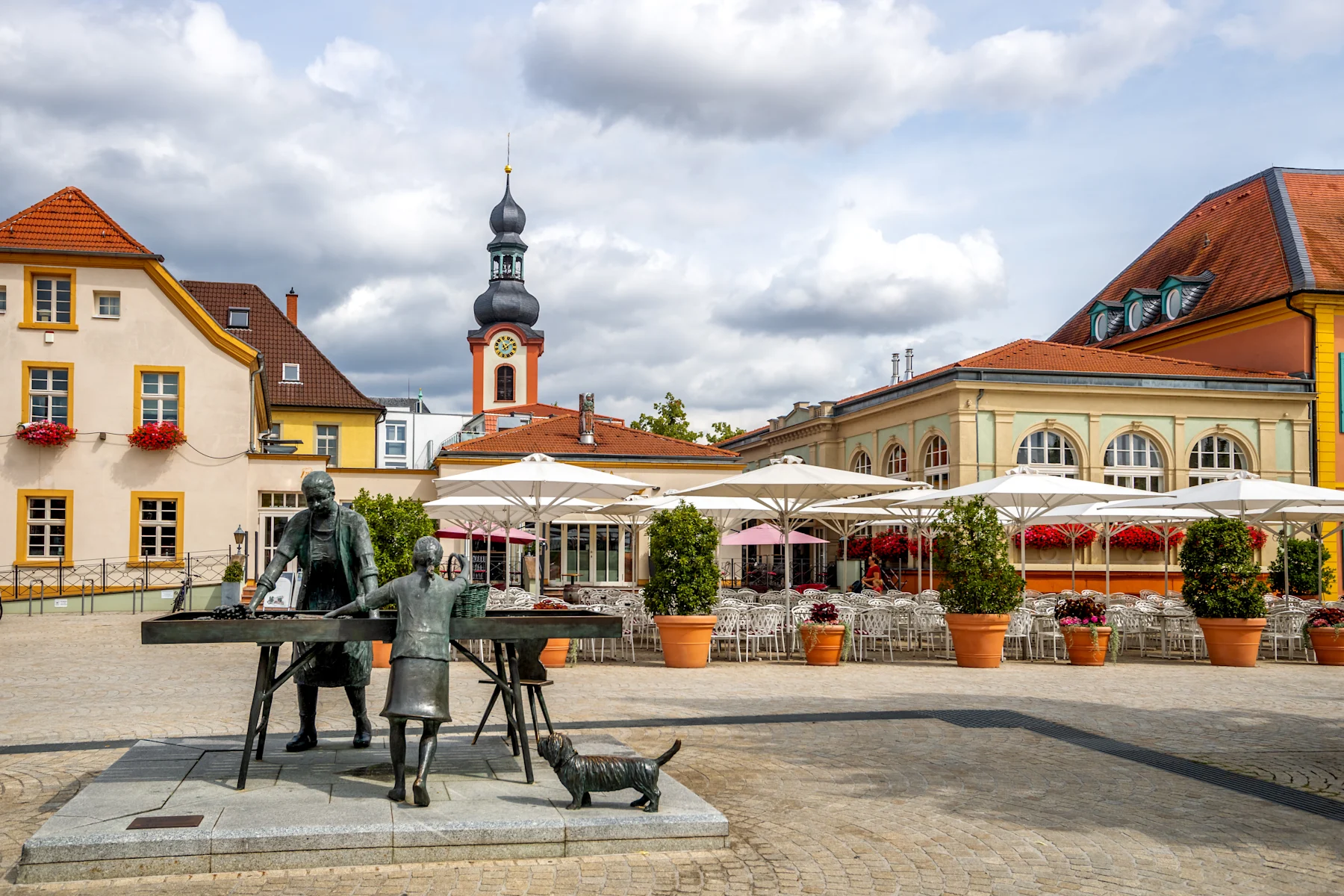 Platz mit Skulpturengruppe, aufgestellten Sonnenschirmen und bunten Häusern im Hintergrund.