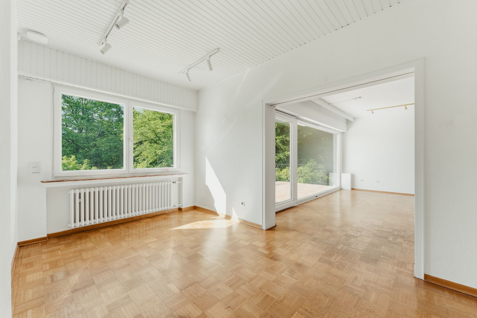 Heller Raum mit Parkettboden, weißen Wänden und großen Fenstern mit Blick ins Grüne.