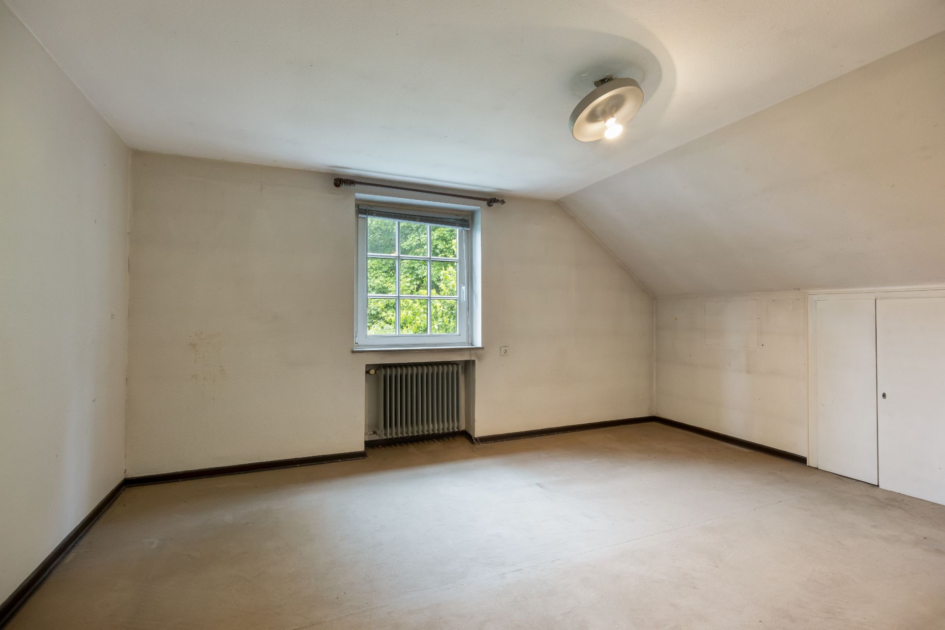 Leerer Raum mit Fenster, Heizkörper und Deckenleuchte, Wand mit kleiner Tür.