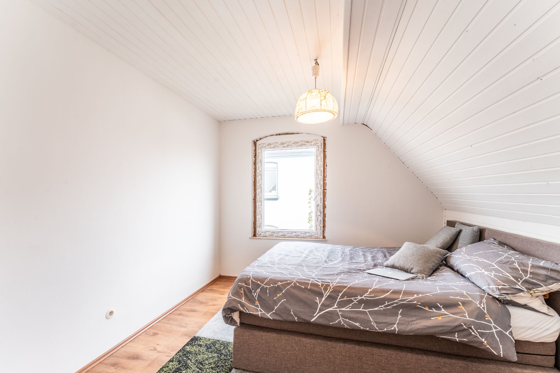 Helles Schlafzimmer mit Doppelbett unter Schräge, Fenster und Lampe an der Decke.