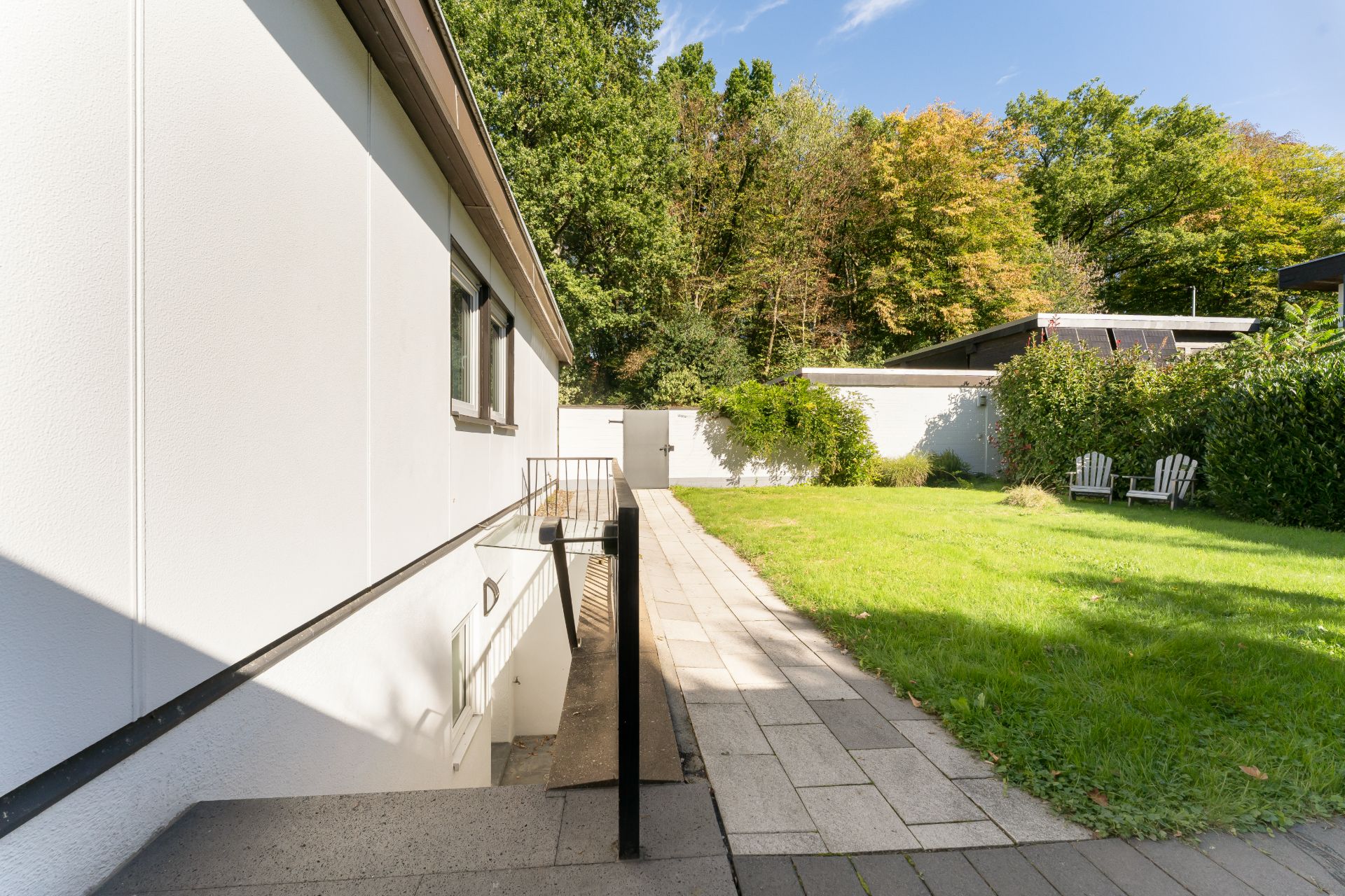 Gepflegter Garten mit Rasen, Terrasse, Weg und zwei weißen Gartenstühlen neben einem modernen Haus.
