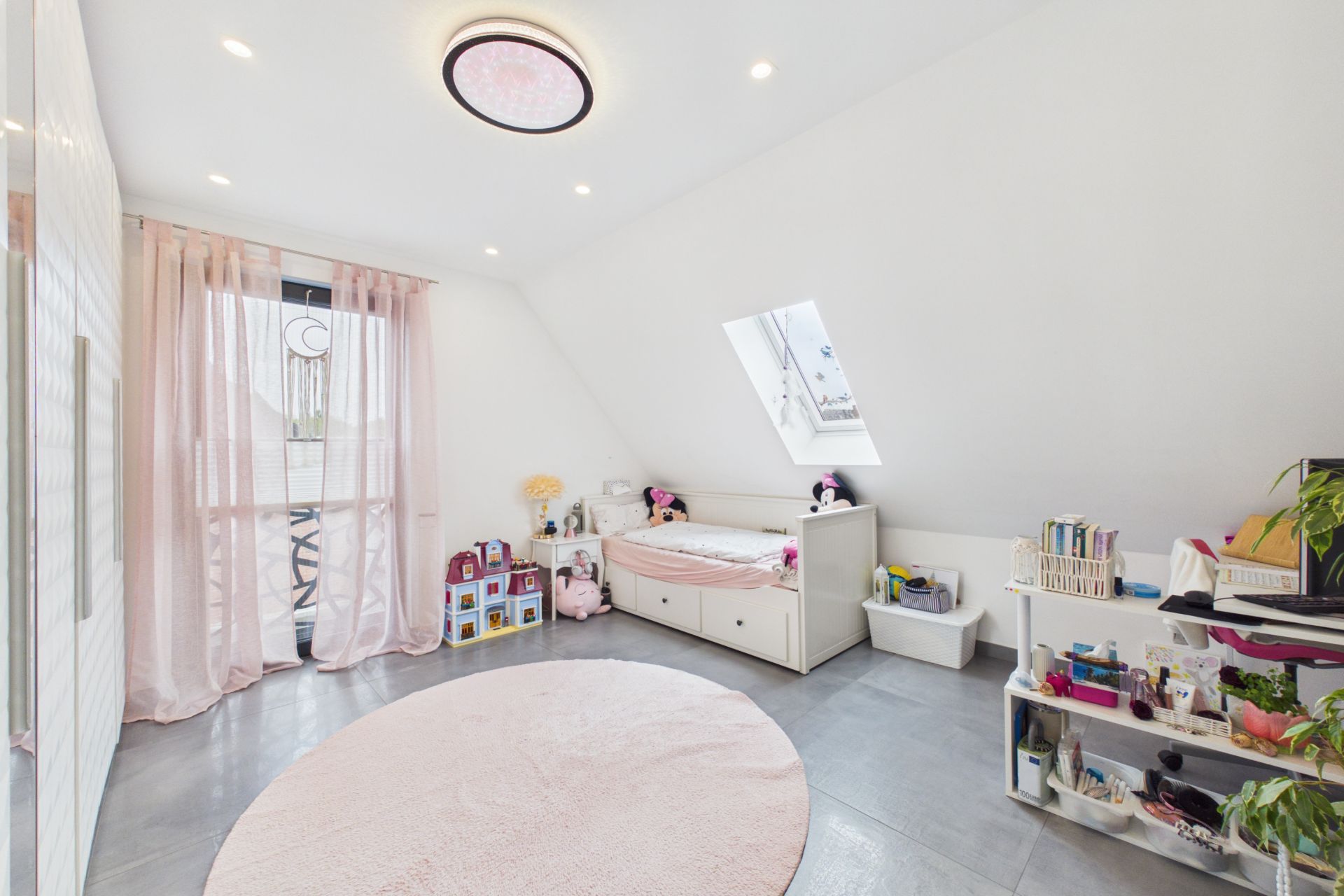 Helles Kinderzimmer mit rundem Teppich, Einzelbett, Regalen und Spielzeug unter einem Dachfenster.