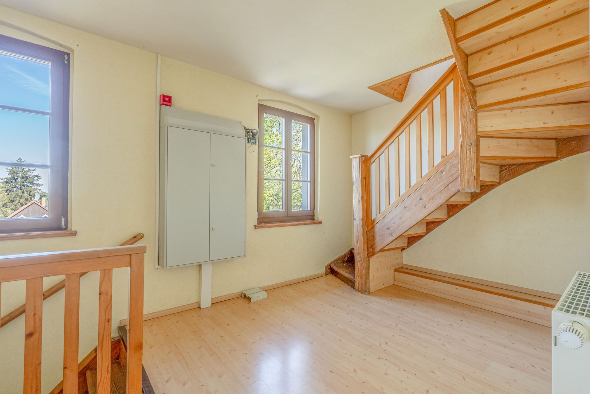 Heller Flur mit Holztreppe, zwei Fenstern und Heizkörper, Parkettboden.