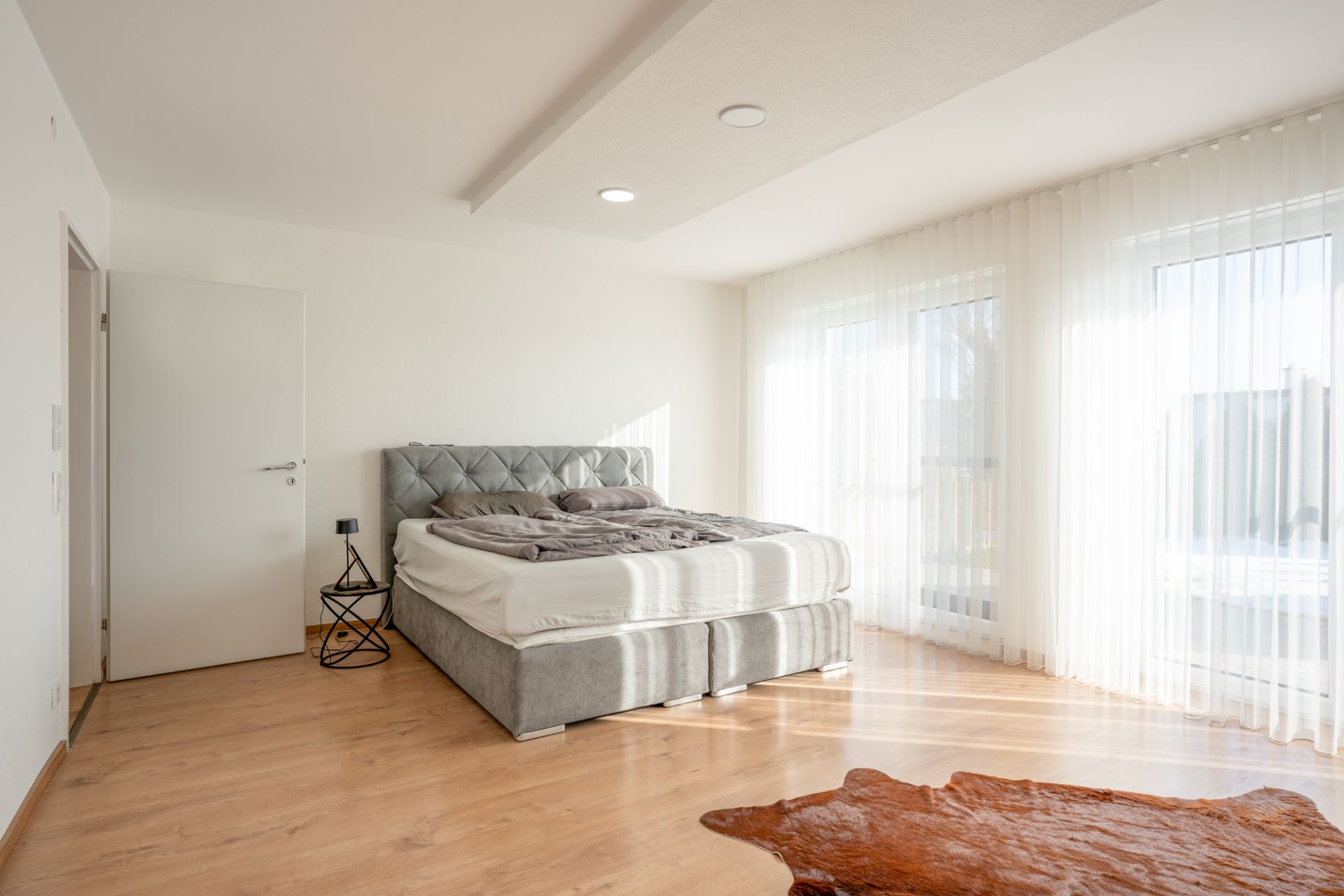 Helles Schlafzimmer mit Doppelbett, Nachttisch und großen Fenstern mit weißen Vorhängen.