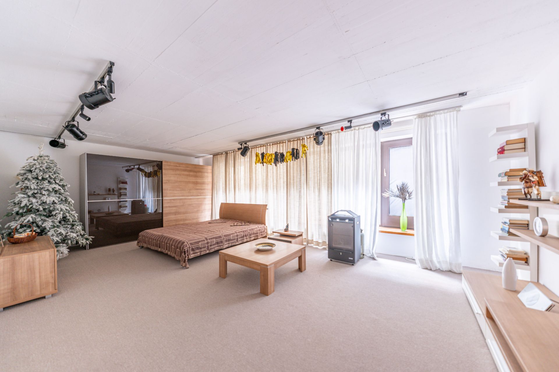 Helles Schlafzimmer mit Doppelbett, Couchtisch, Bücherregalen und einem großen Fenster mit Vorhängen.