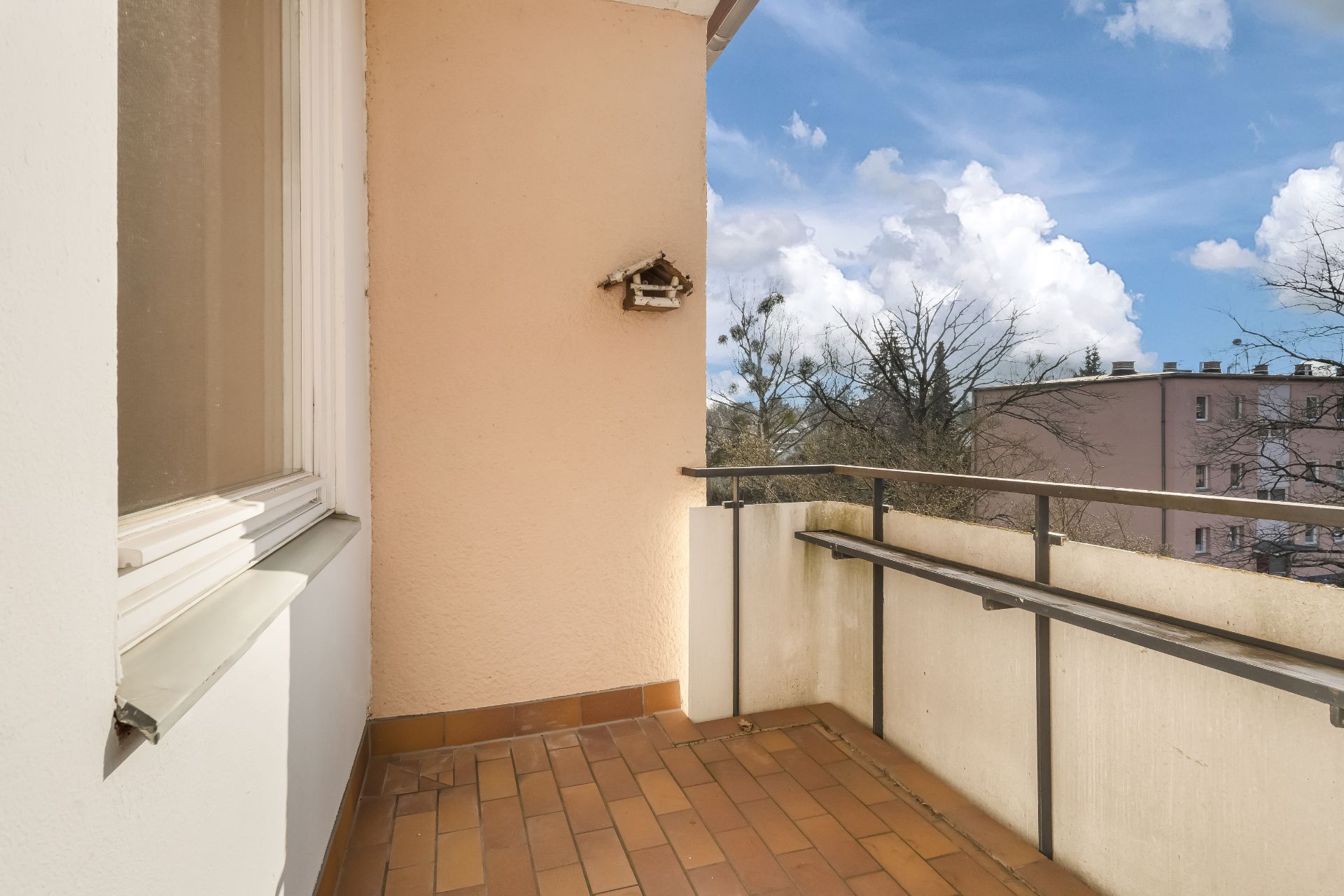Balkon mit braunen Fliesen, Geländer und Ausblick auf Bäume und Wohngebäude.