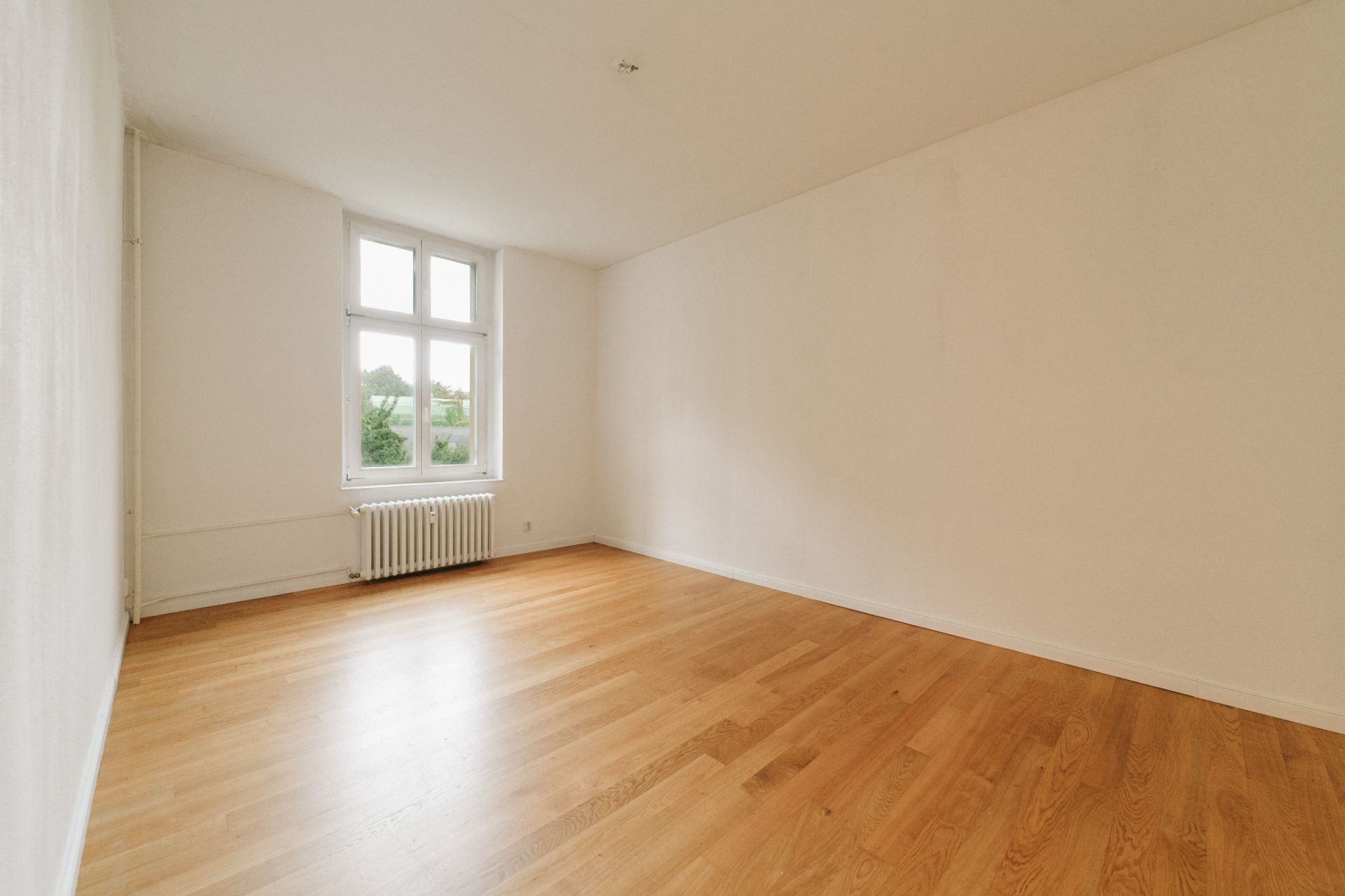 Leeres Zimmer mit Holzboden, weiß gestrichenen Wänden und großem Fenster.