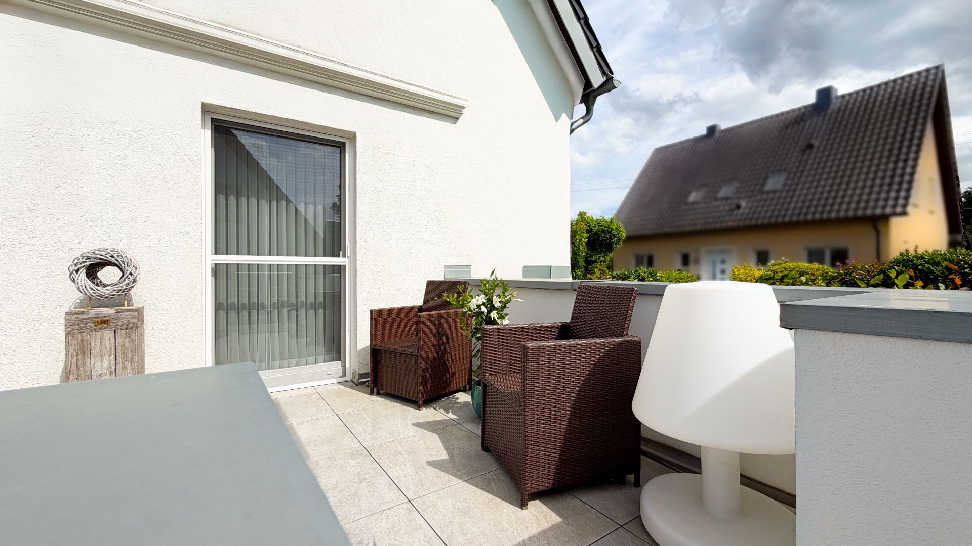 Möblierter Balkon mit Korbstühlen, Beistelltisch und moderner Lampe, angrenzend an ein Wohnhaus.