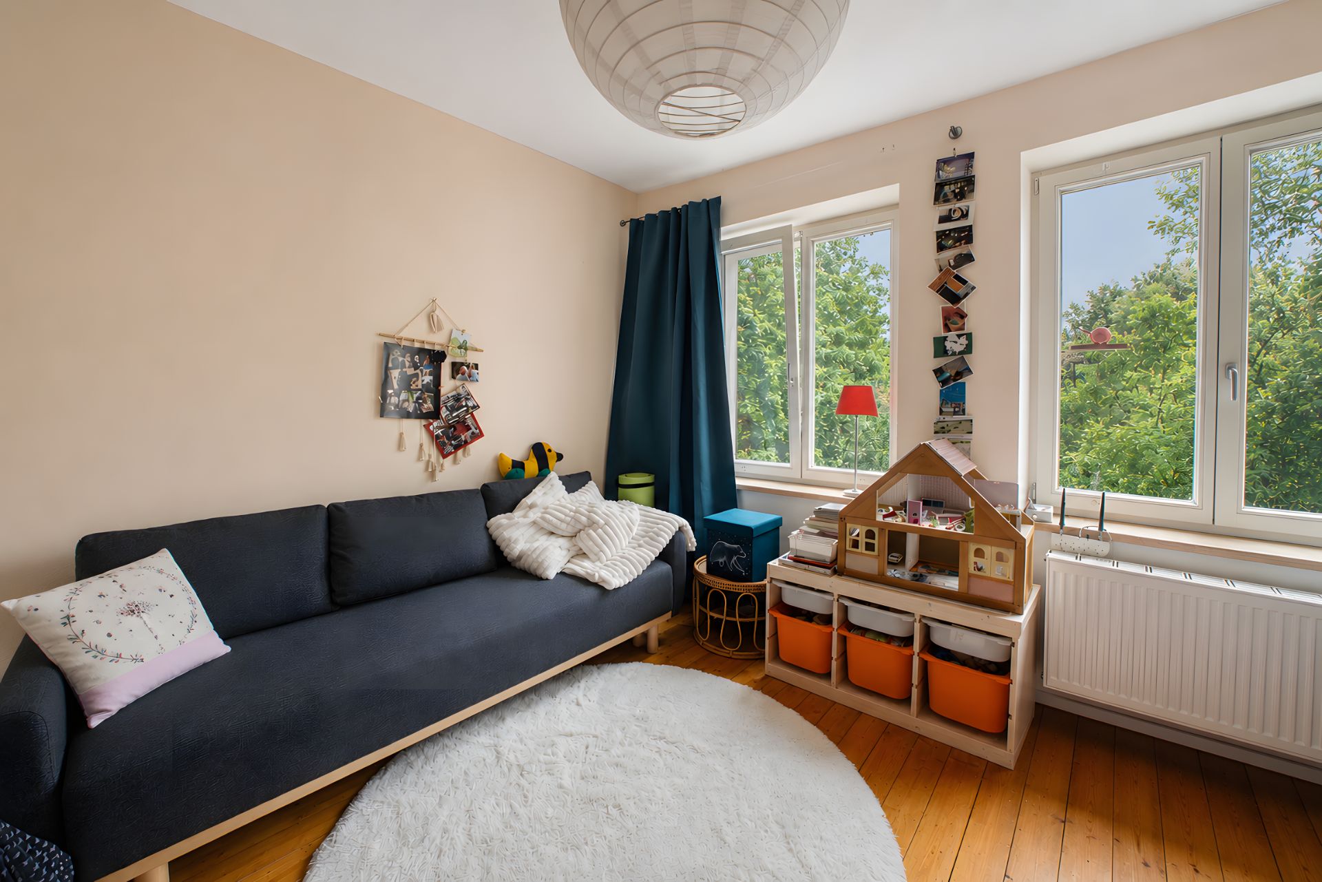 Helles Kinderzimmer mit dunkelgrauem Sofa, Spielzeugregal und zwei großen Fenstern.