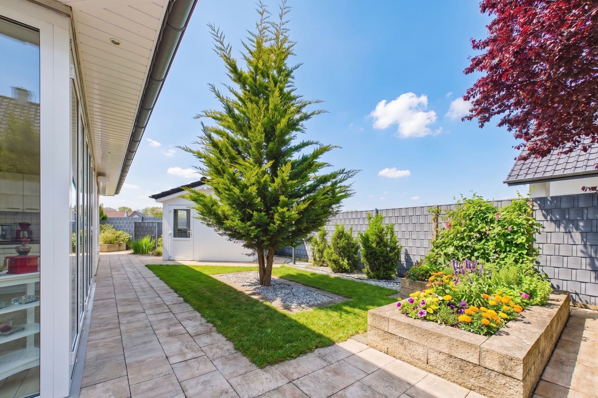Gepflegter Garten mit Baum, Blumenbeet und gepflastertem Weg neben einem Wohnhaus.