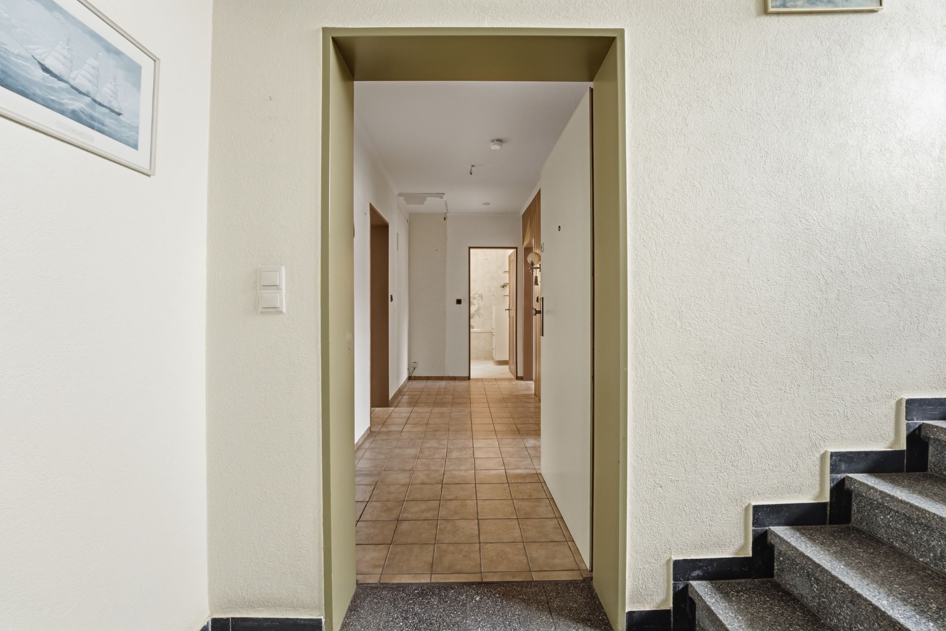 Blick in einen Flur mit Fliesenboden, weißen Wänden und einer Treppe rechts im Vordergrund.