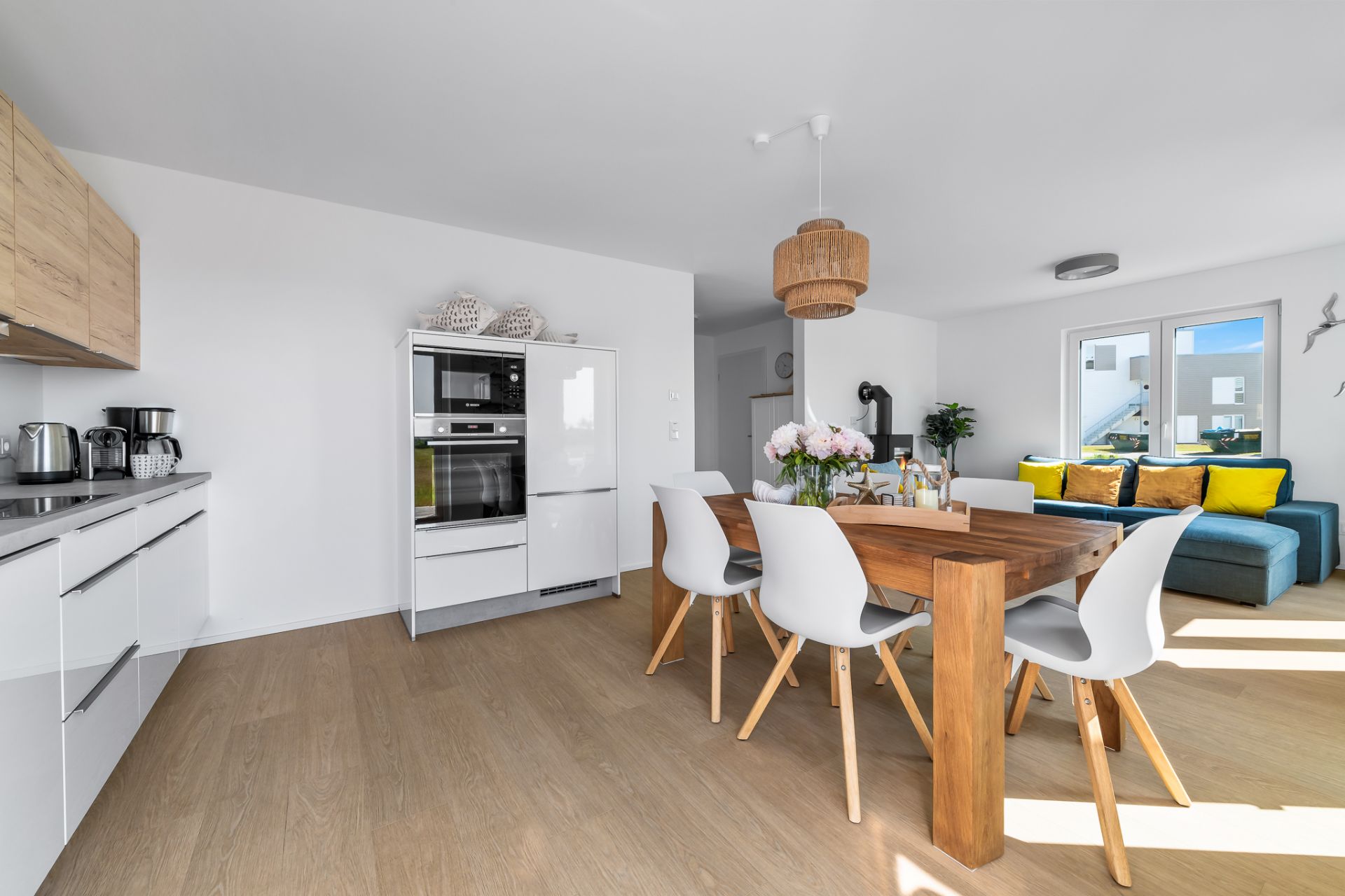 Helle Wohnküche mit Holztisch, weißen Stühlen und Sofa mit bunten Kissen am Fenster.