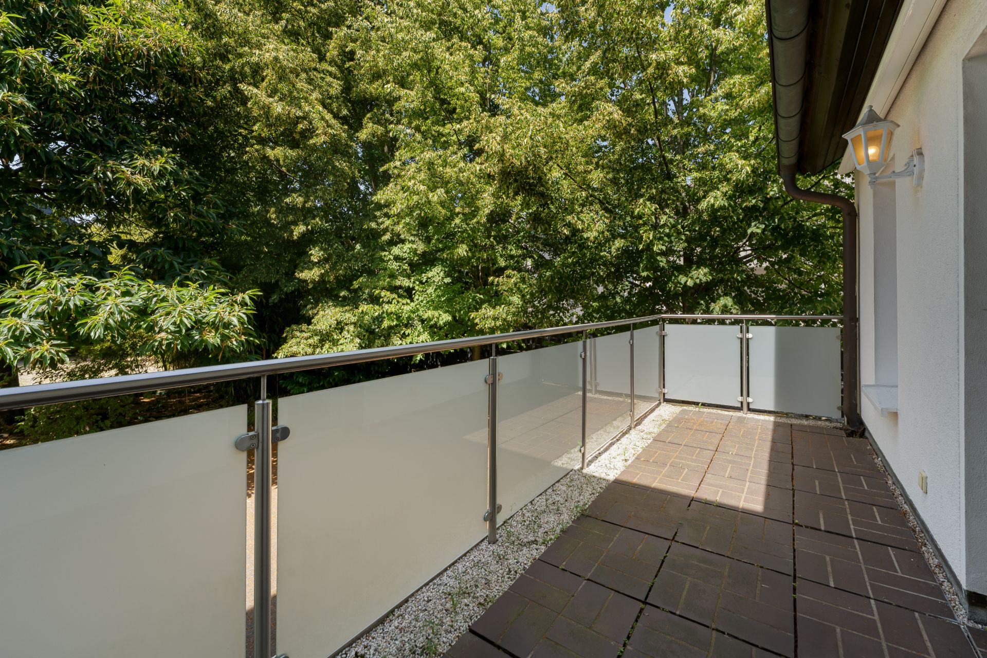 Balkon mit Glasgeländer und braunen Bodenfliesen, umgeben von grünen Bäumen.