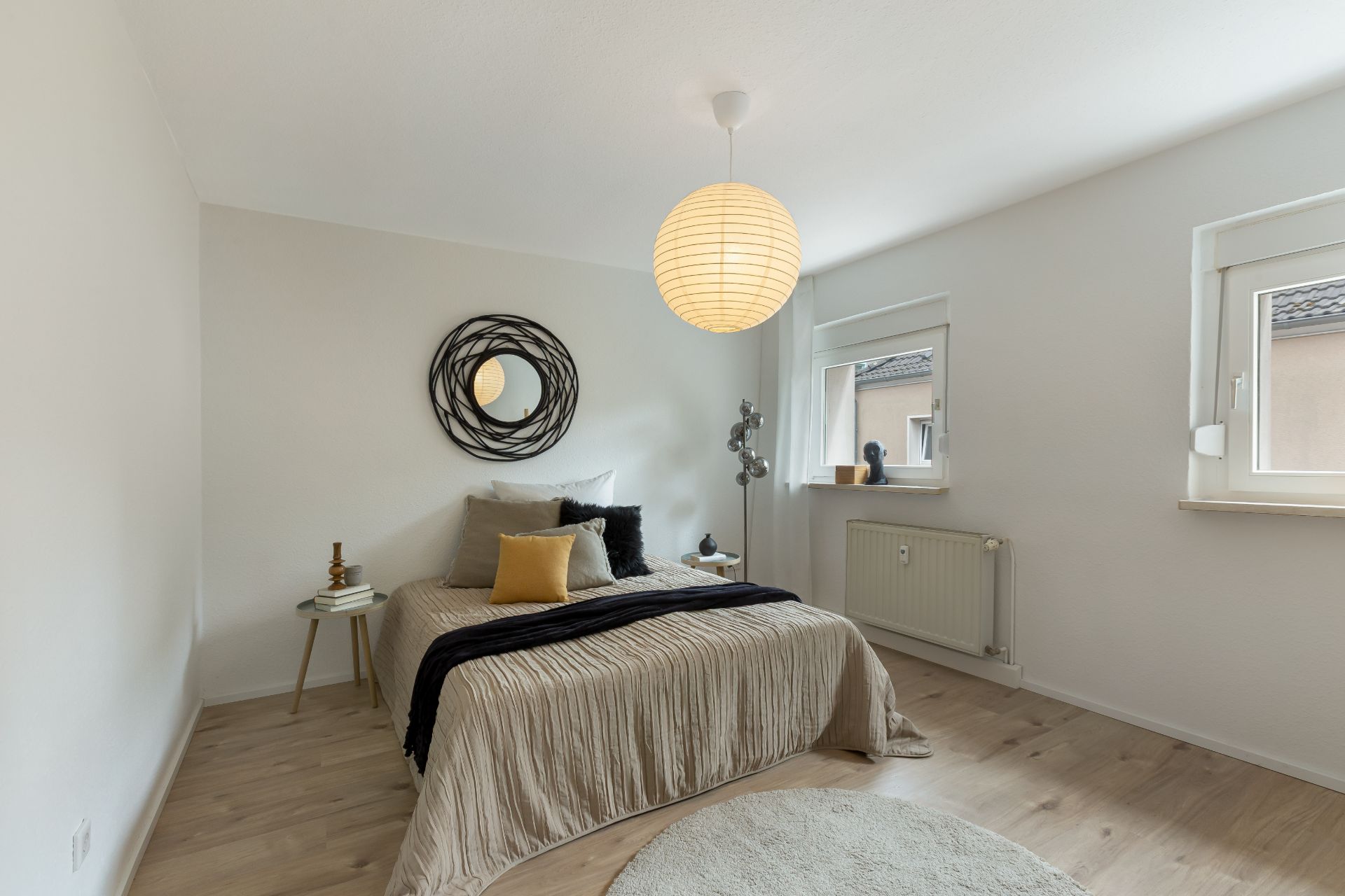 Helles Schlafzimmer mit Doppelbett, runder Lampe, Spiegel und zwei Fenstern.