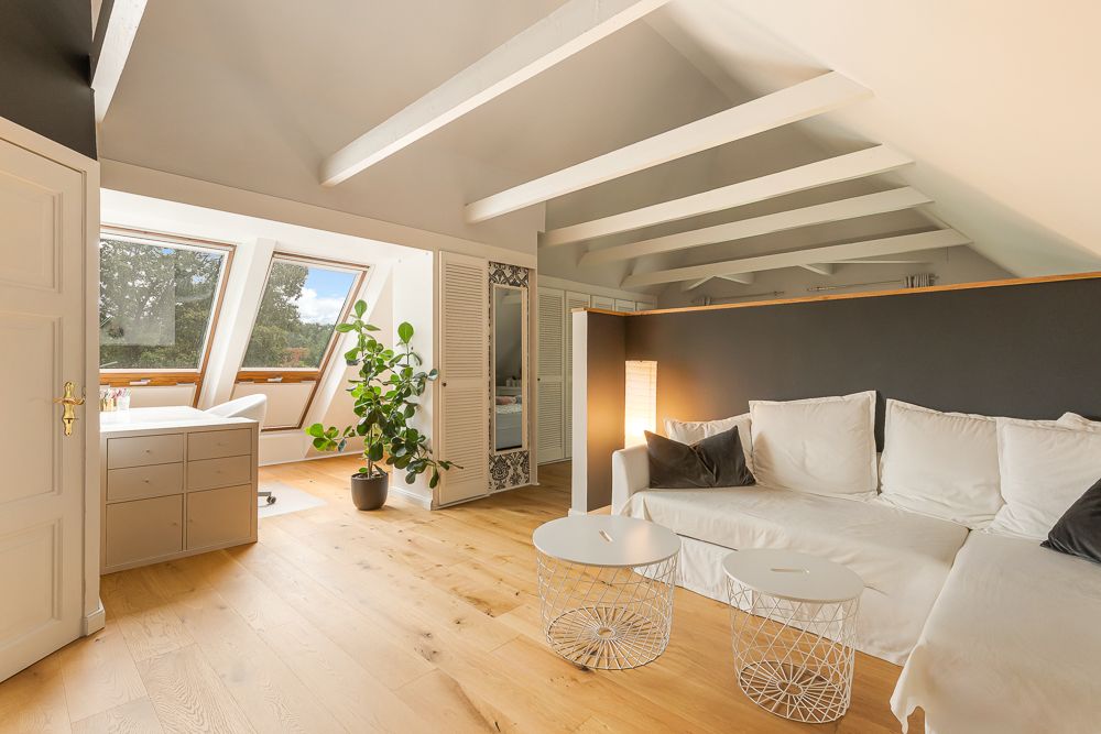 Helles Wohnzimmer mit weißem Sofa, zwei runden Tischen, großer Pflanze und Schreibtisch vor Dachfenstern.