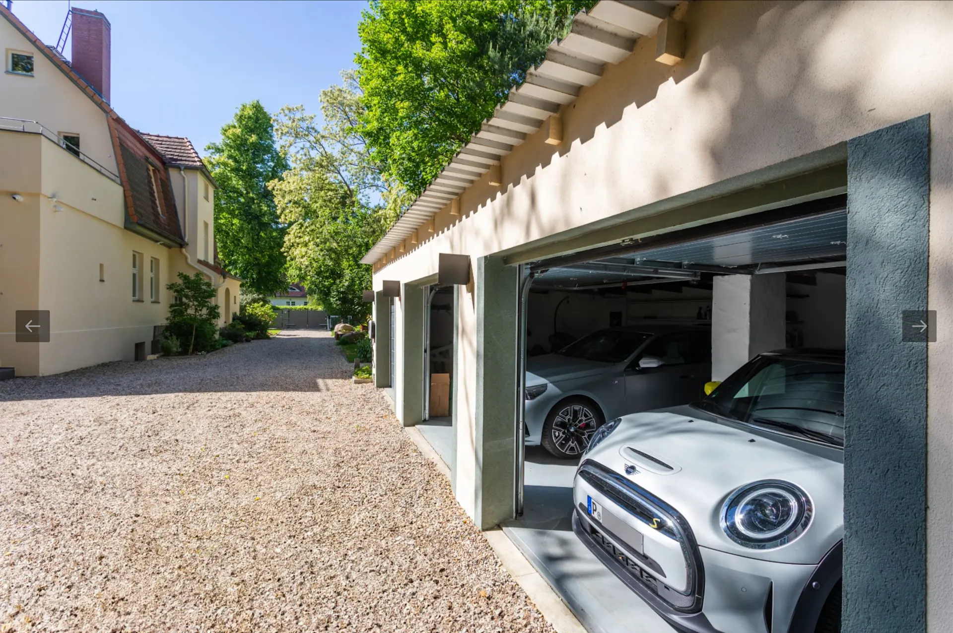 Zwei Autos parken in einer offenen Garage neben einem Wohnhaus mit Kiesauffahrt.