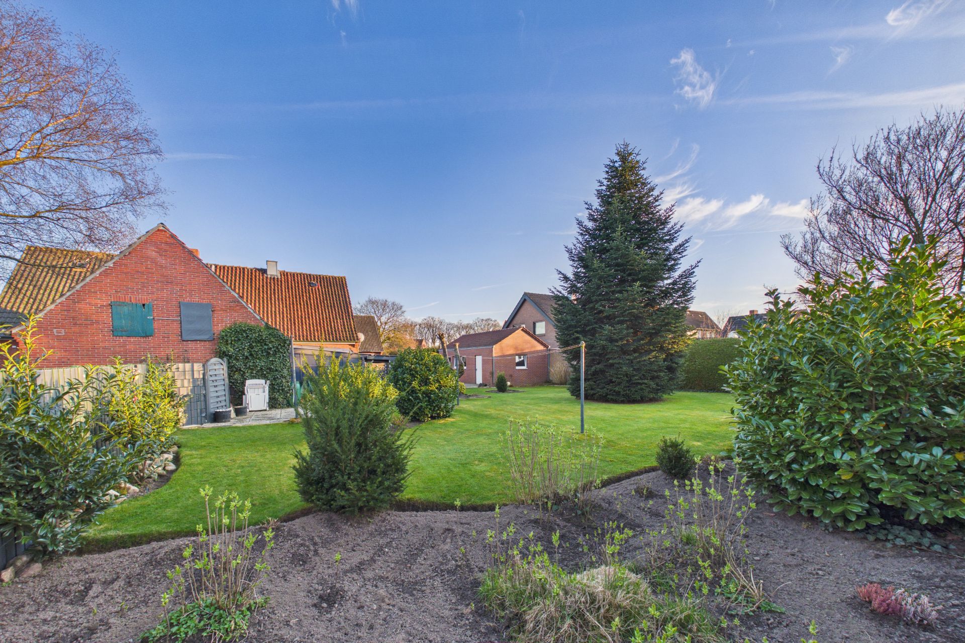 Großer Garten mit Rasen, Sträuchern, Bäumen und einem Backsteinhaus im Hintergrund.
