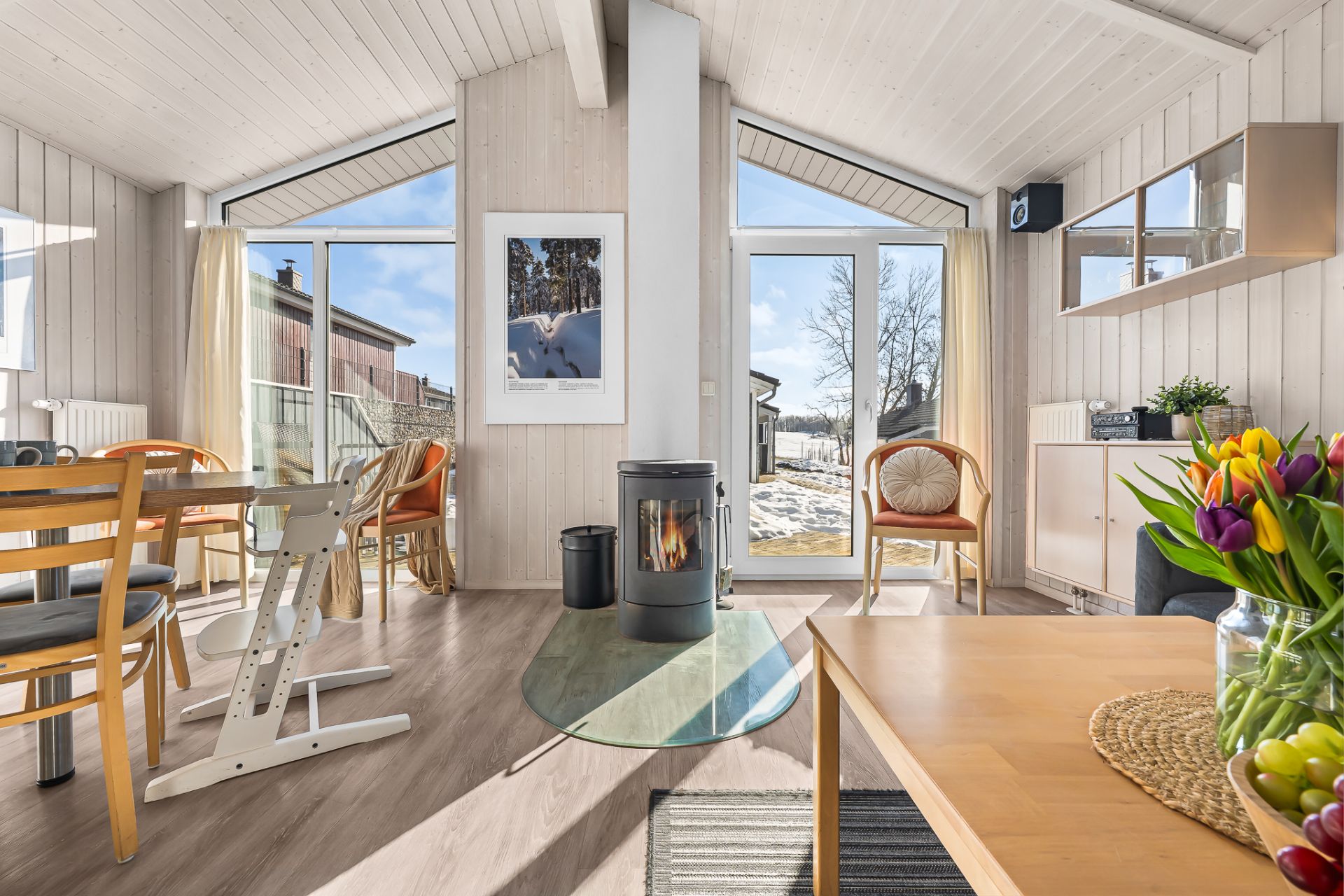 Helles Wohnzimmer mit Kamin, Holzmöbeln, Esstisch und großen Fenstern mit Blick nach draußen.