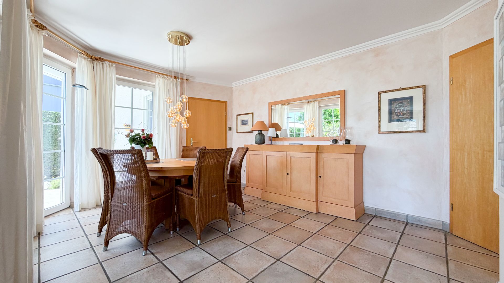 Esszimmer mit rundem Holztisch, sechs Korbstühlen, Sideboard, Spiegel und Fliesenboden.