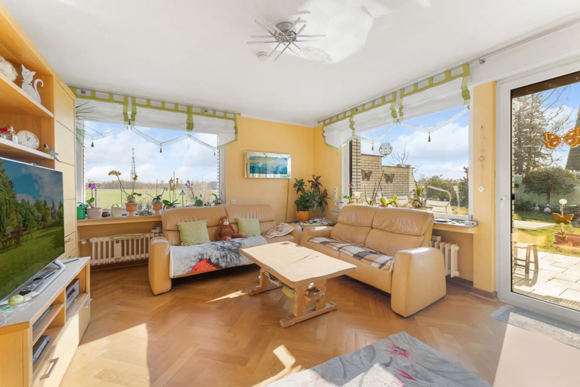 Helles Wohnzimmer mit zwei Ledersofas, Couchtisch, Fernseher und großen Fenstern mit Blick in den Garten.