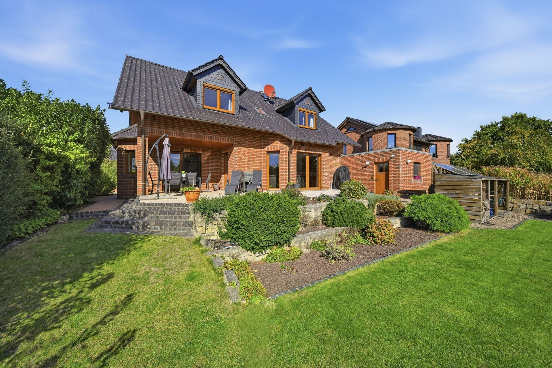 Backsteinwohnhaus mit Terrasse, Gartenmöbeln und gepflegtem Garten bei sonnigem Wetter.
