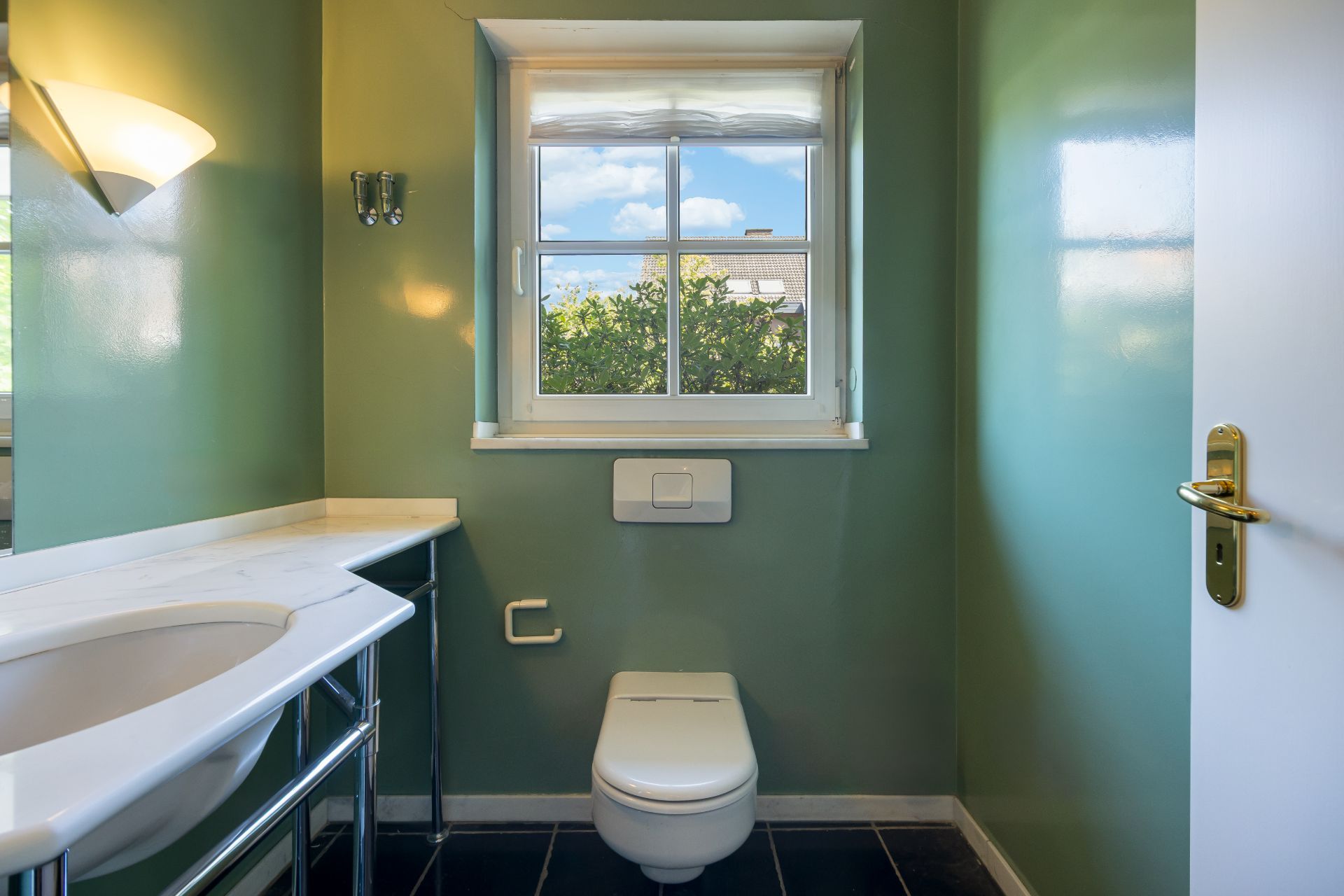 Kleines Badezimmer mit grün gestrichenen Wänden, WC, Waschbecken und Fenster mit Blick nach draußen.
