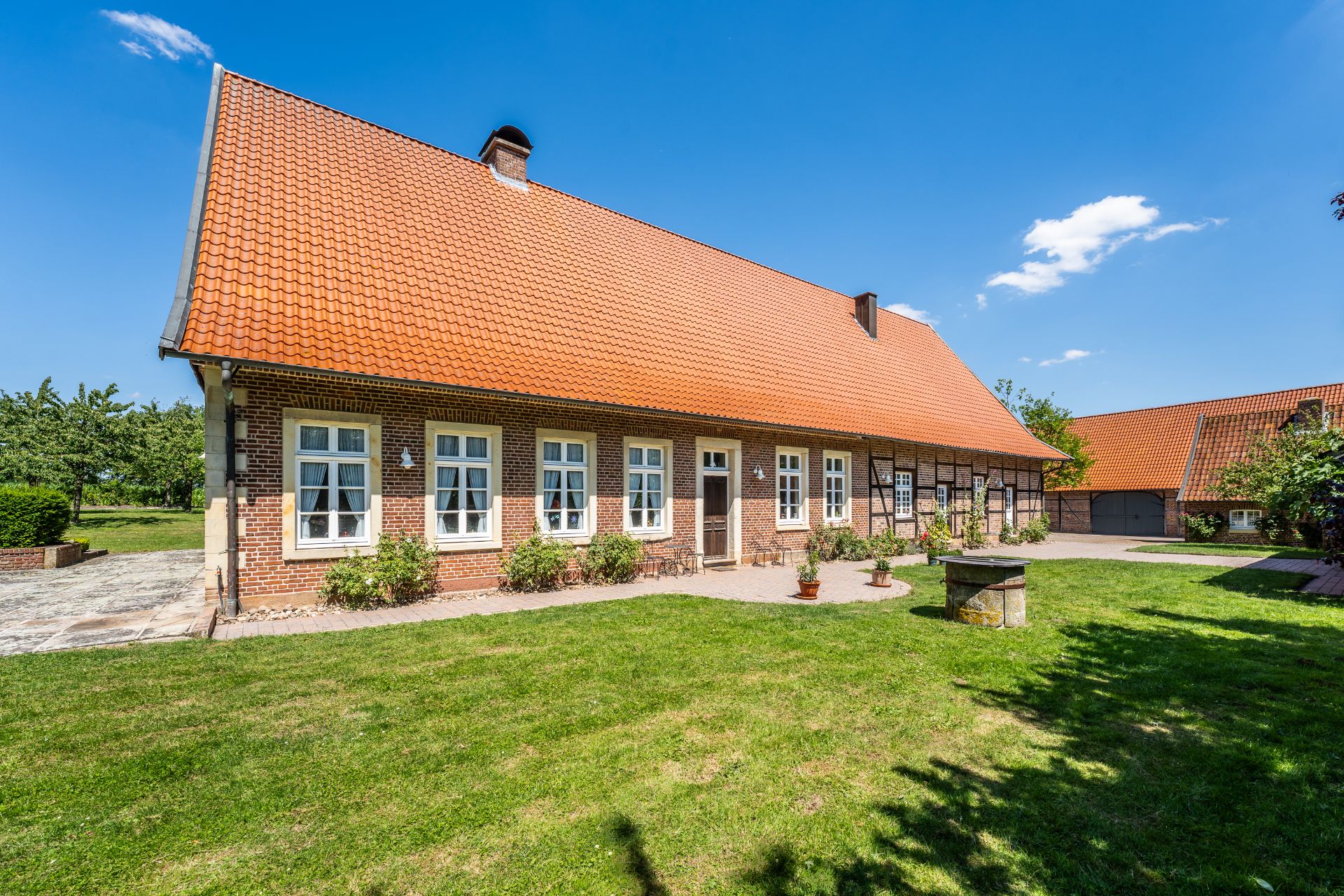 Backsteinhaus mit rotem Dach, mehreren Fenstern und grünem Garten bei sonnigem Wetter.