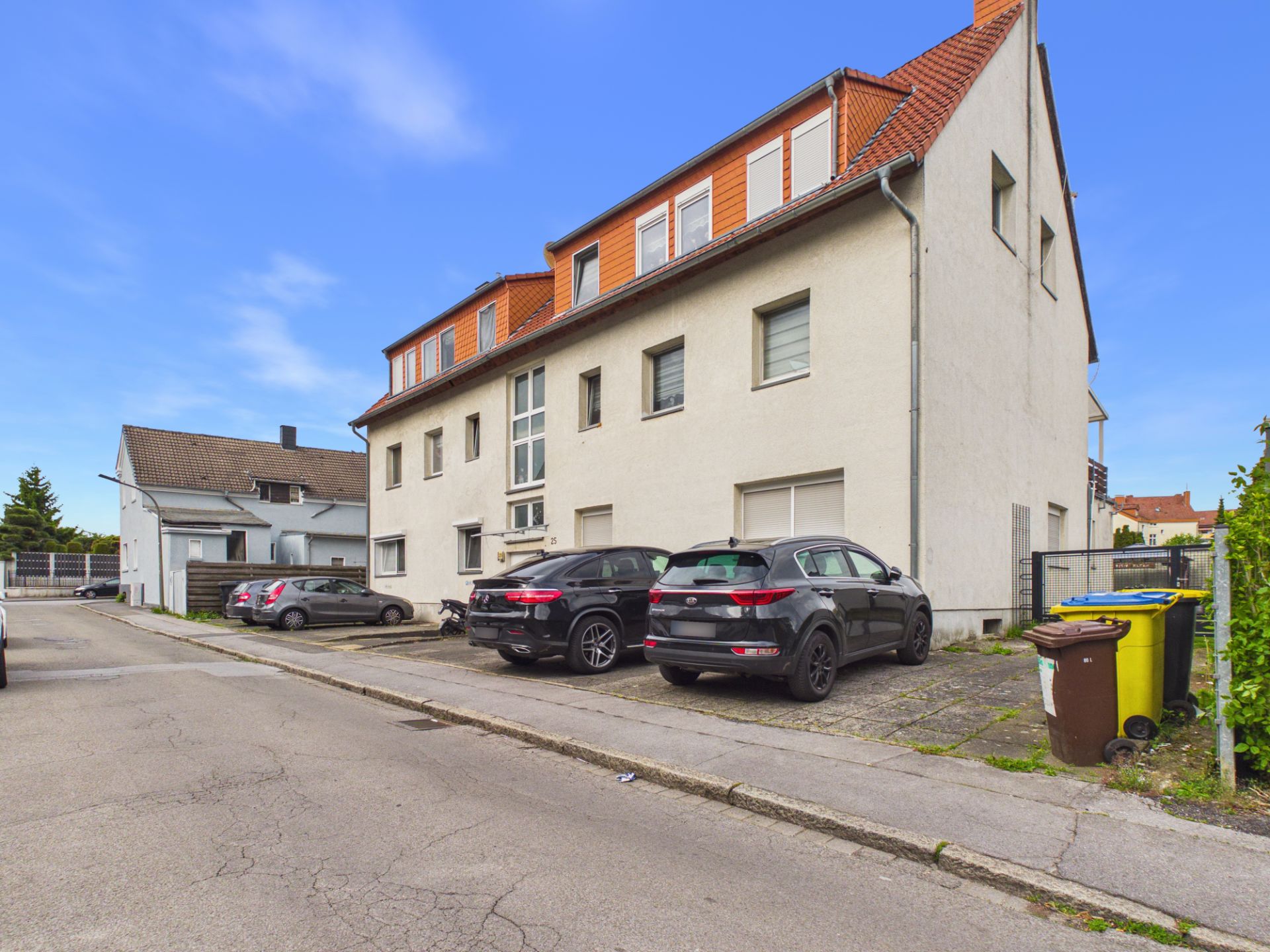 Mehrfamilienhaus mit roten Dachziegeln, mehreren geparkten Autos und Mülltonnen am Straßenrand.