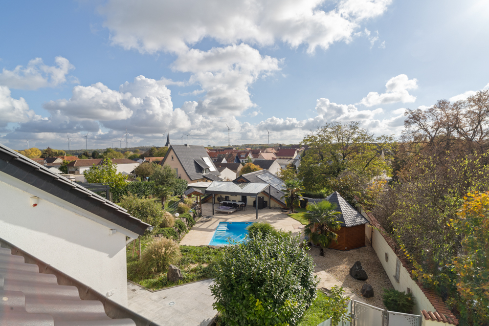 Blick auf einen Garten mit Pool, Terrasse und umliegenden Bäumen, Häuser im Hintergrund.