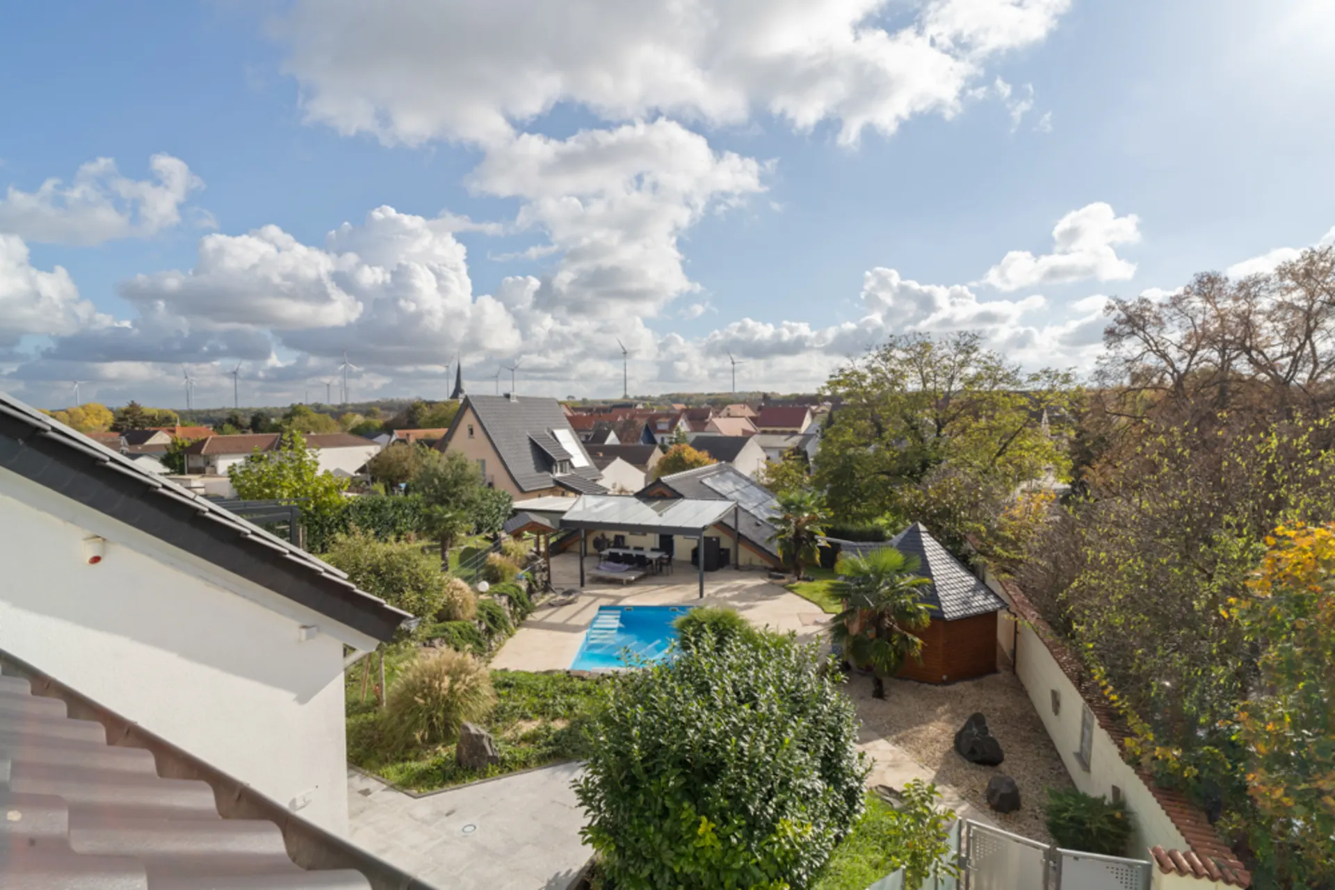 Blick auf einen Garten mit Pool, Terrasse und umliegenden Bäumen, Häuser im Hintergrund.