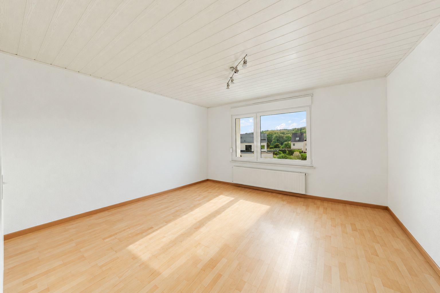 Leerer Raum mit hellem Holzboden, weißer Decke und großem Fenster.