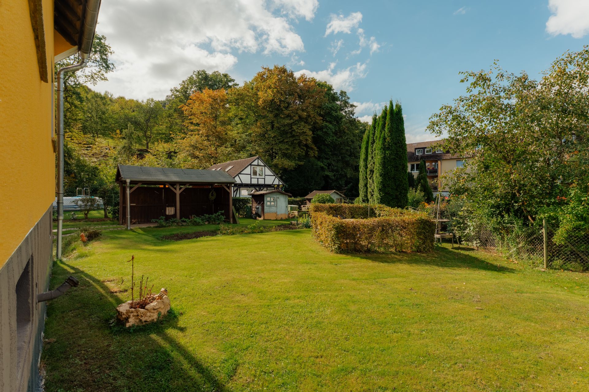 Grüner Garten mit Rasenfläche, Hecke, Bäumen und einem kleinen Holzgebäude im Hintergrund.