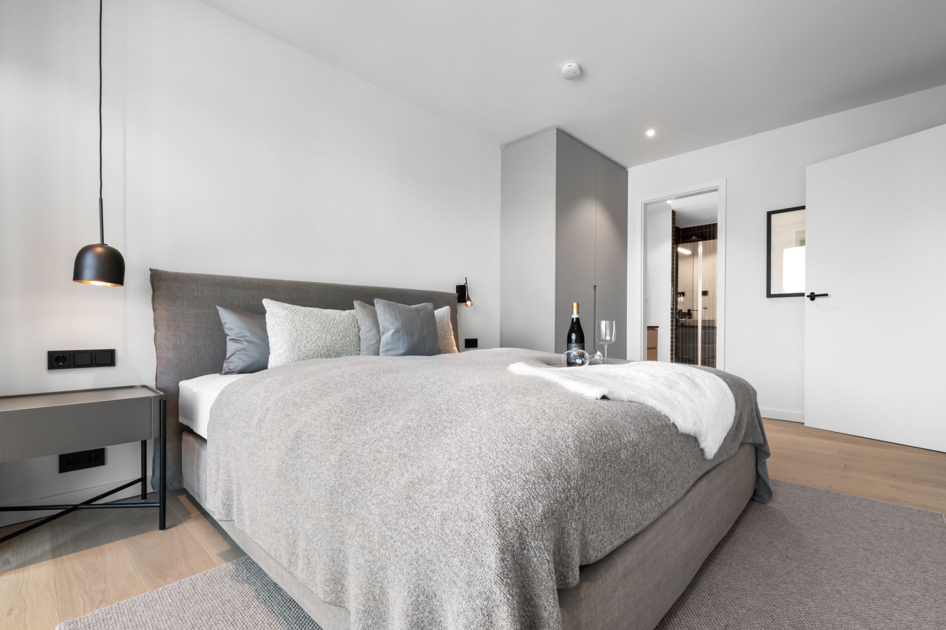 Modernes Schlafzimmer mit grauem Bett, Nachttisch, Flaschenwein und zwei Gläsern auf dem Bett.