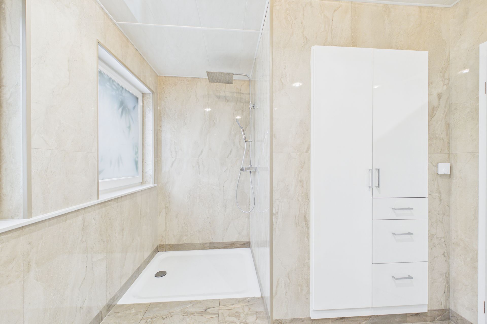 Helles Badezimmer mit großer Dusche, Fenster und weißem Schrank.