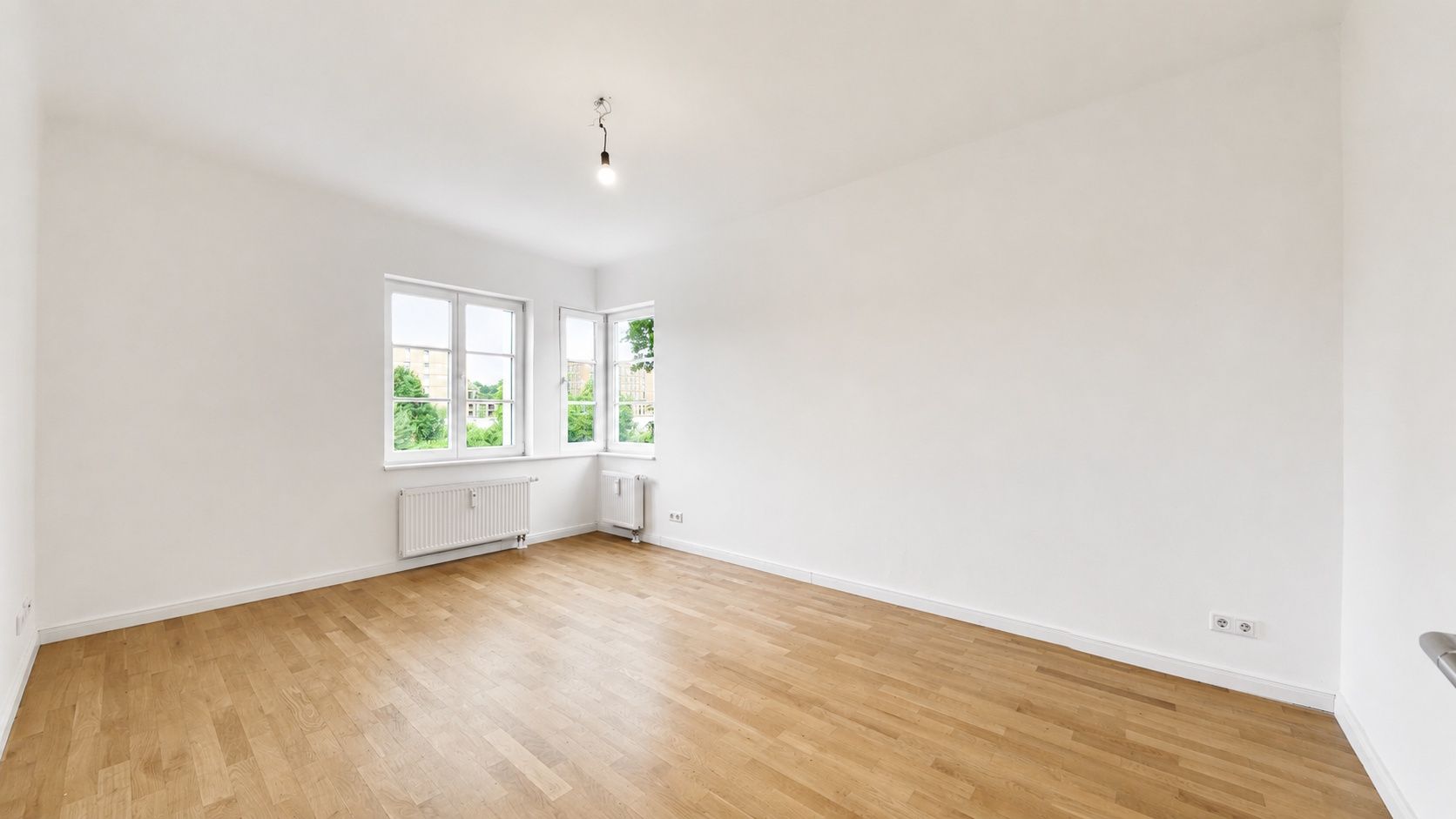 Helles, leerstehendes Zimmer mit Holzfußboden, weißen Wänden und zwei Fenstern.