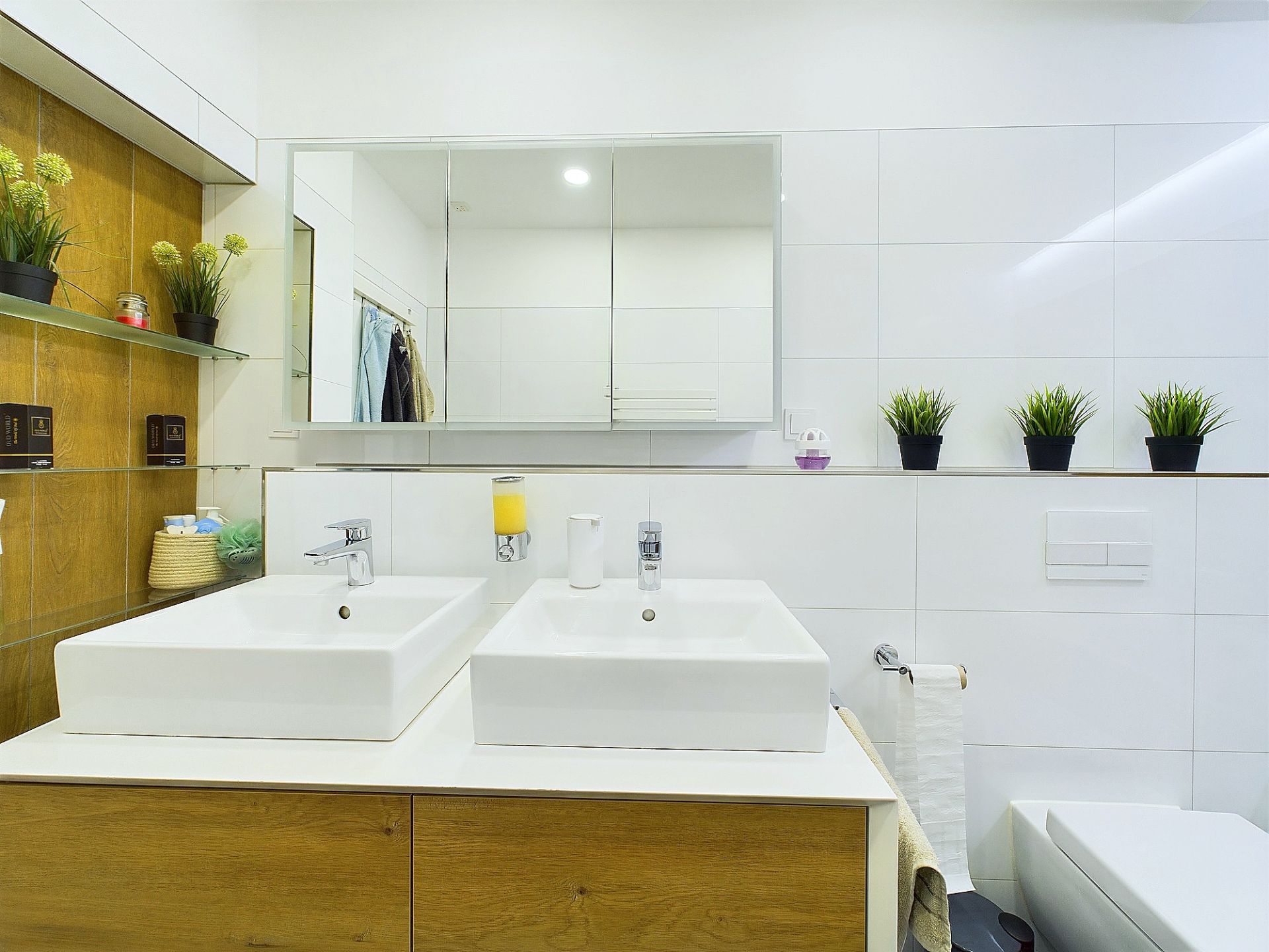 Modernes Badezimmer mit zwei Waschbecken, großem Spiegel und Dekopflanzen auf weißen Fliesen.