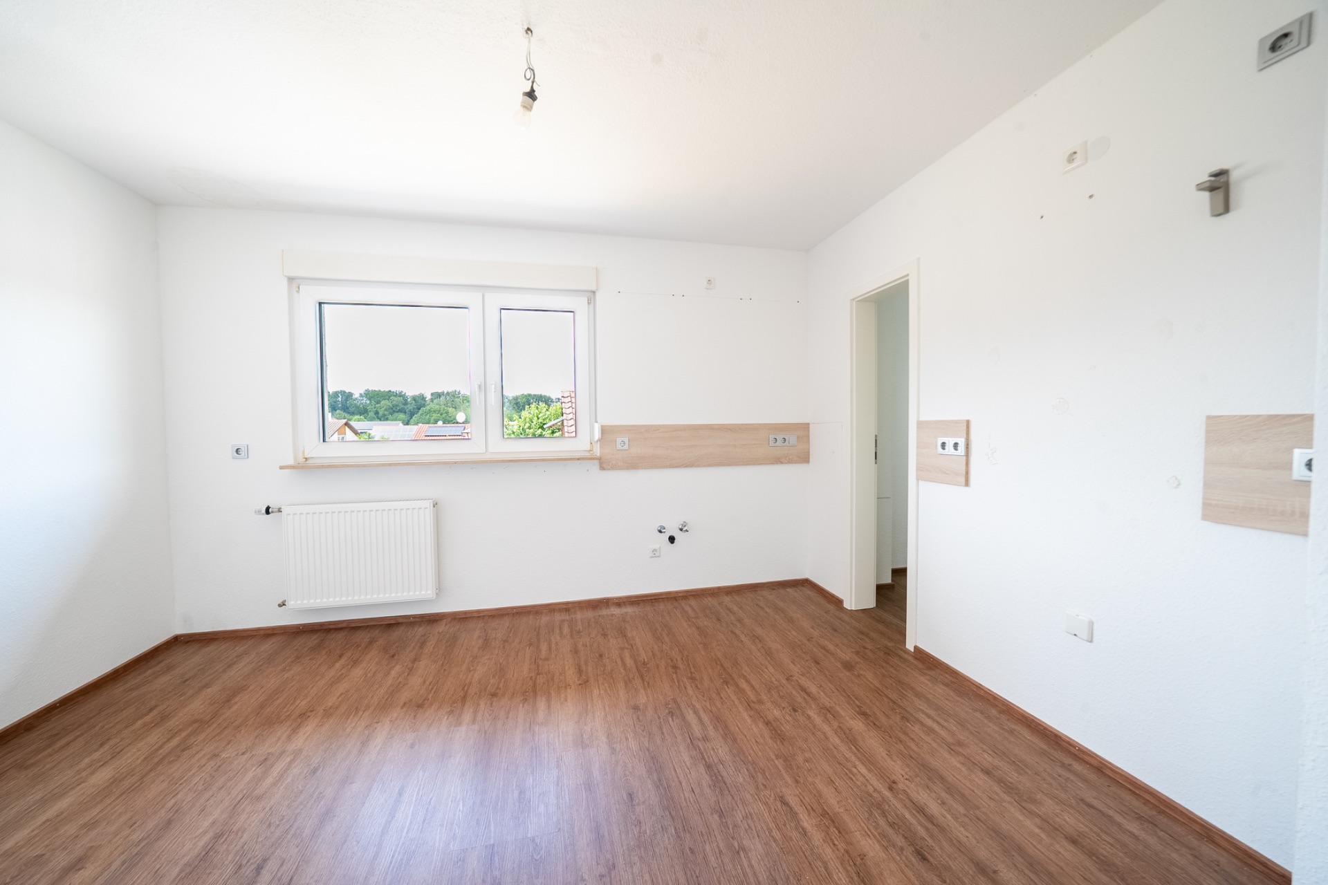 Leerer Raum mit weißer Wand, großem Fenster, Holzfußboden und Heizkörper.