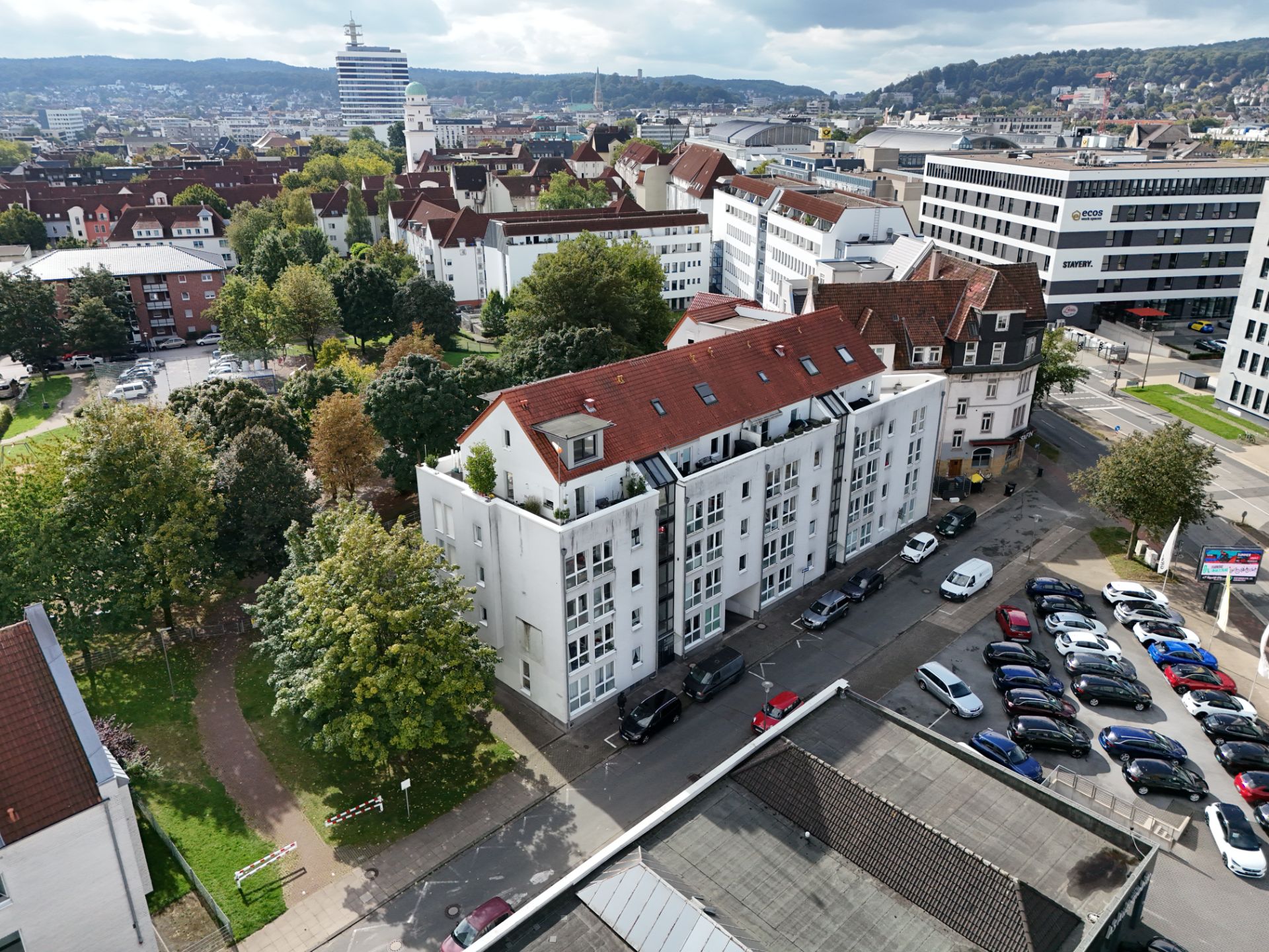 Wohnhaus mit rotem Dach, Balkonen und geparkten Autos, umgeben von Bäumen in einer Stadt.