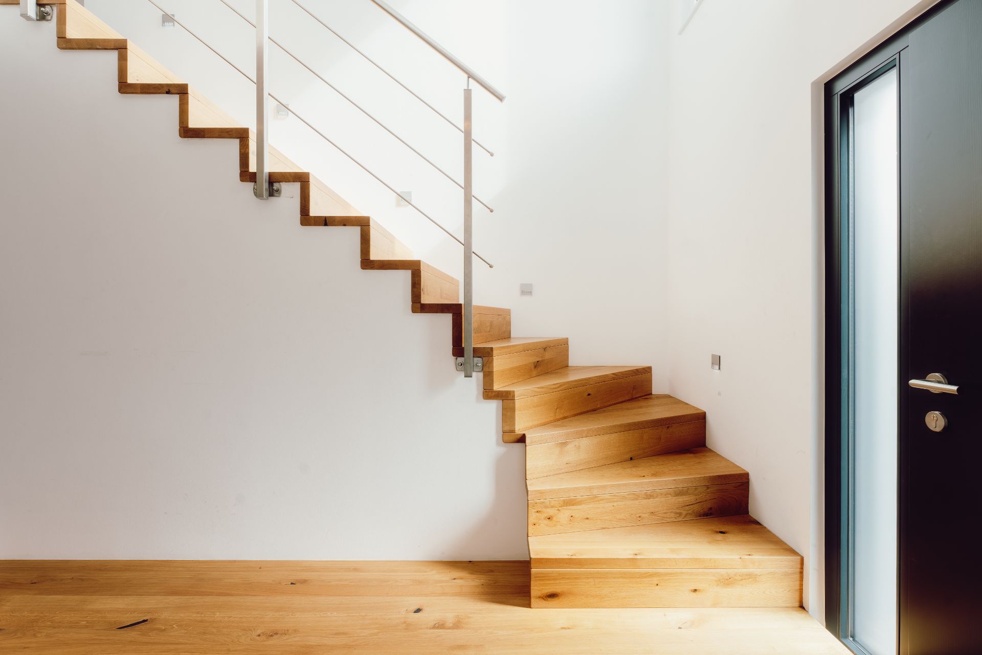 Moderne Holztreppe mit Metallgeländer in einem hellen Flur neben einer schwarzen Tür.