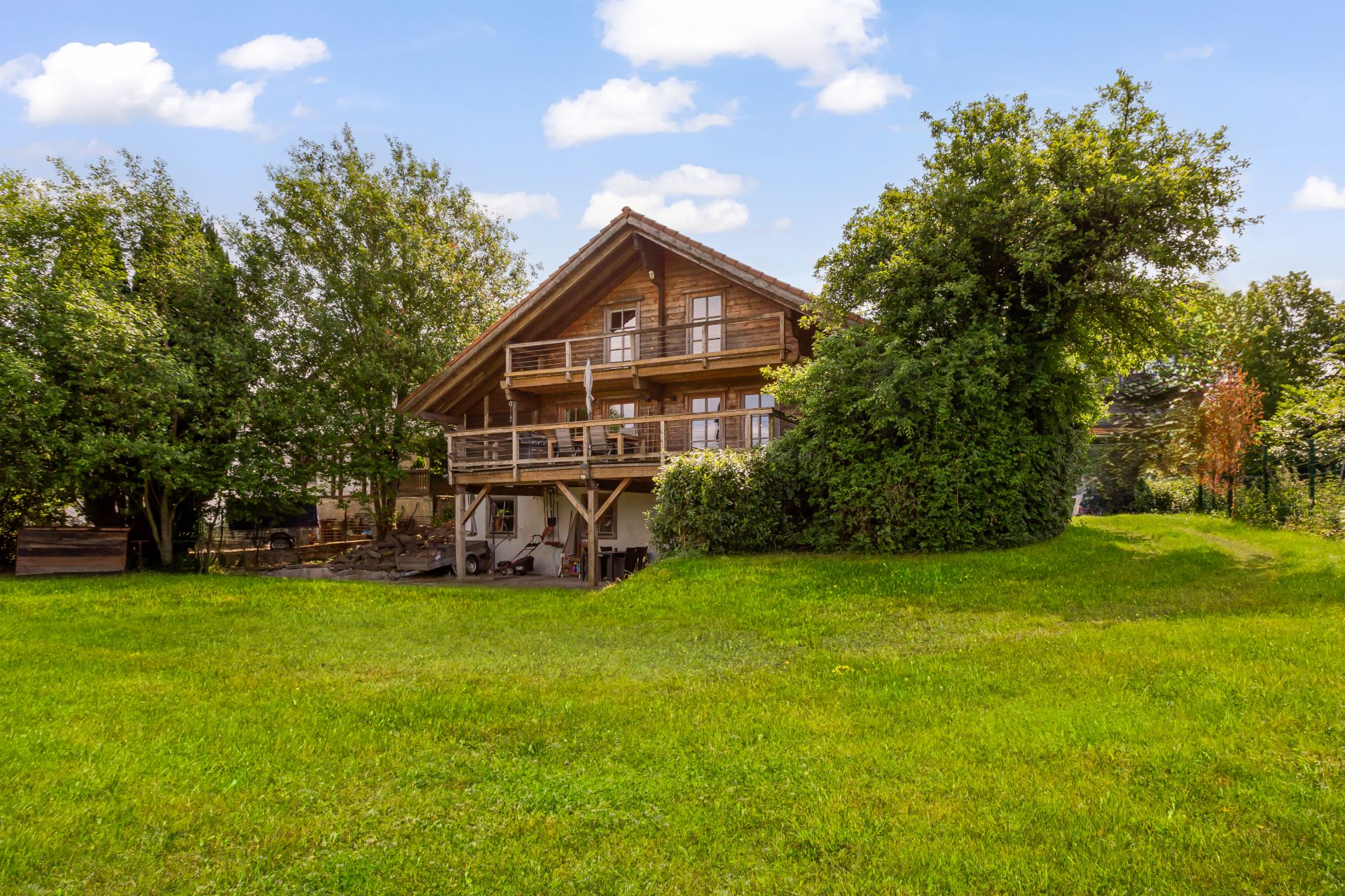 Holzhaus mit Balkon und Garten, umgeben von Bäumen und Rasen bei Tageslicht.