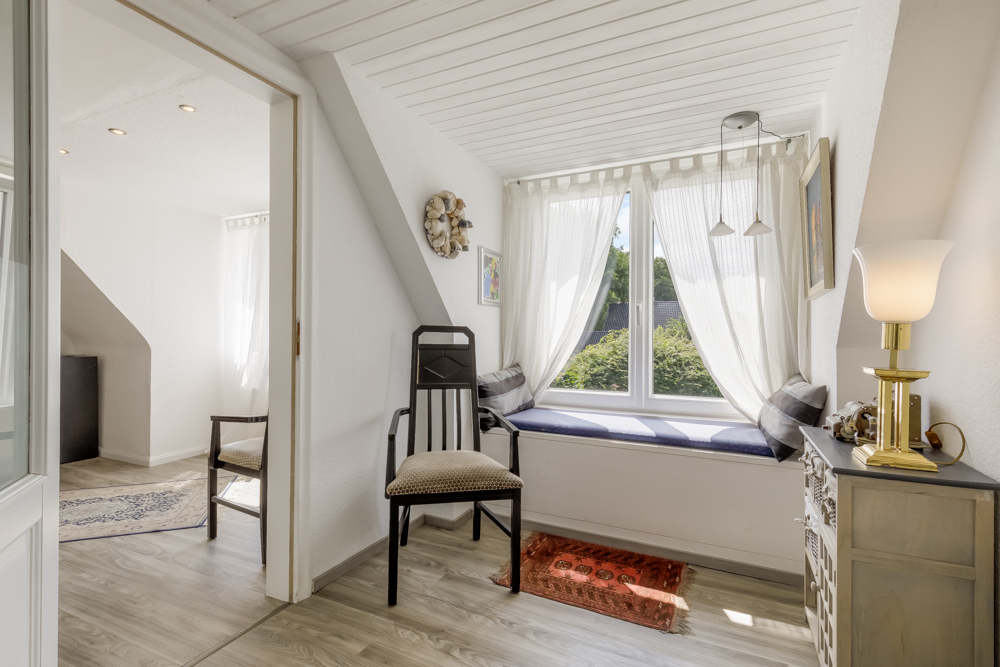 Heller Raum mit Fensterbank, Stuhl, Kommode und Tischlampe, Blick auf begrünte Außenseite.