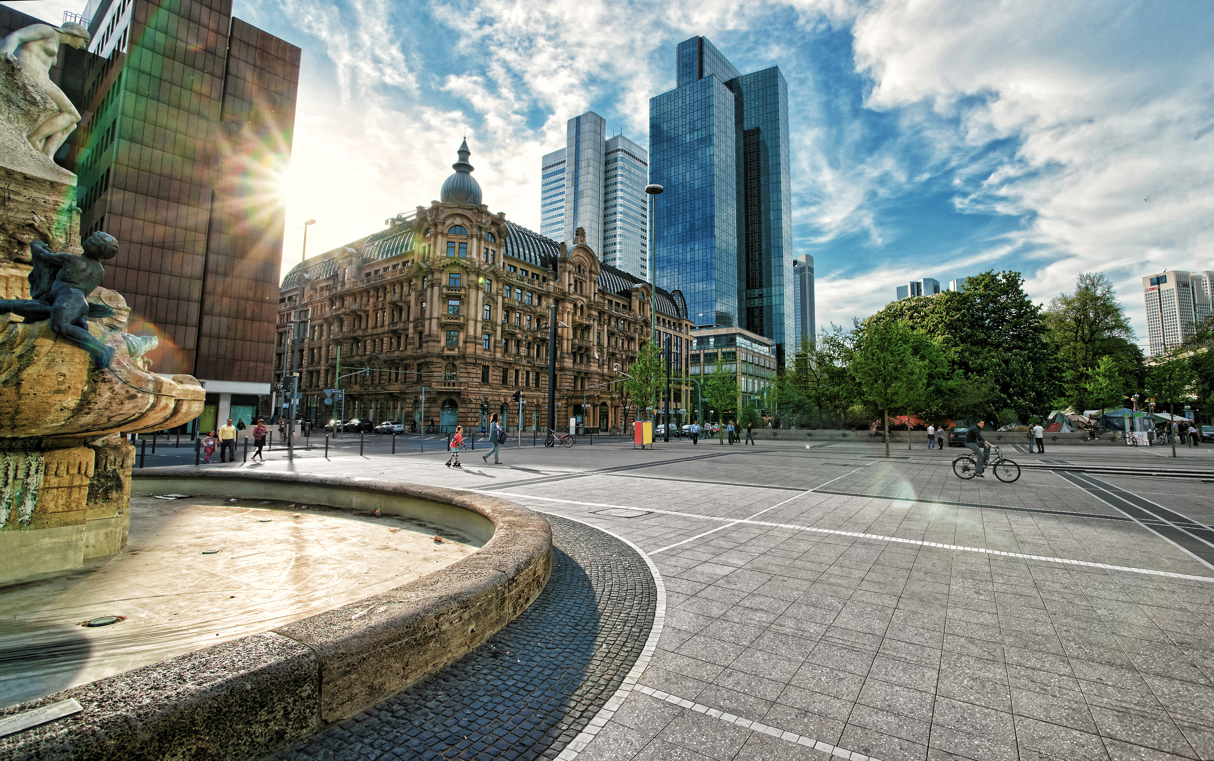 Großer Platz mit einem Brunnen vorne, alten und modernen Gebäuden im Hintergrund.